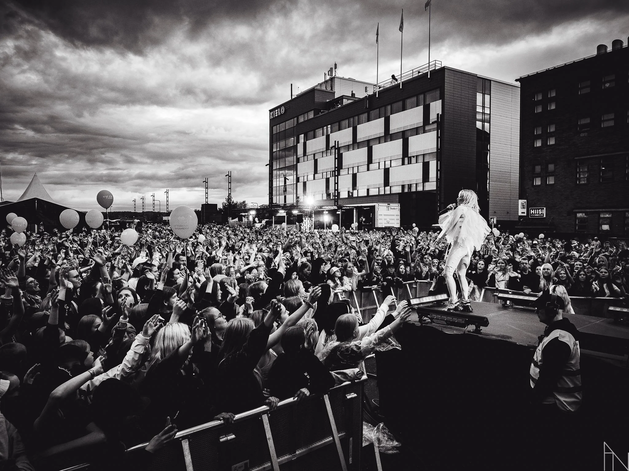 Kuvassa on suuri yleisö tapahtumassa ulkona, jossa esiintyy nainen lavalla. Taustalla on moderni rakennus ja pilvinen taivas.