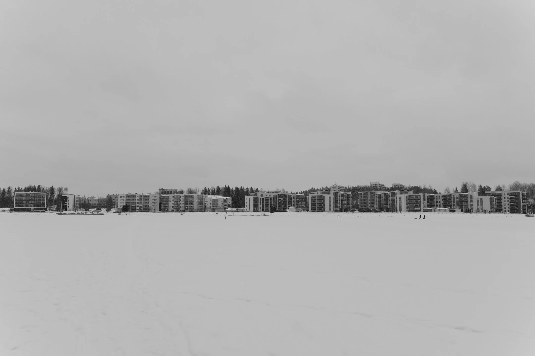 Luminen kaupunkirakennuksia talvisessa maisemassa, lumen peittämällä alueella ja pilviverhossa taivaalla.