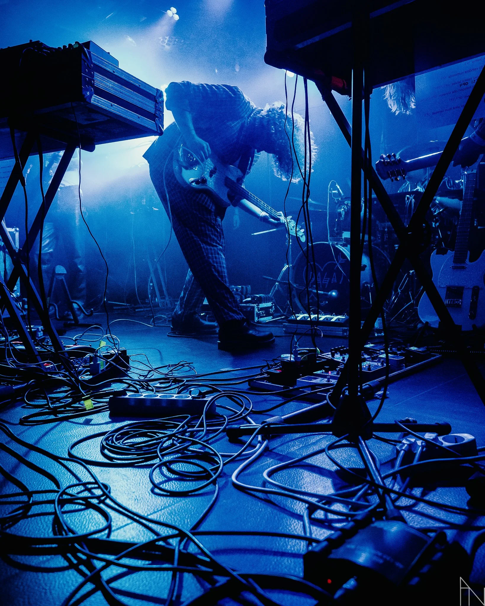 Mies esiintyy lavalla kitaran kanssa, taustalla sininen valaistus ja lähtövaloja, ympärillä monia Kaapeleita ja musiikkilaitteita.