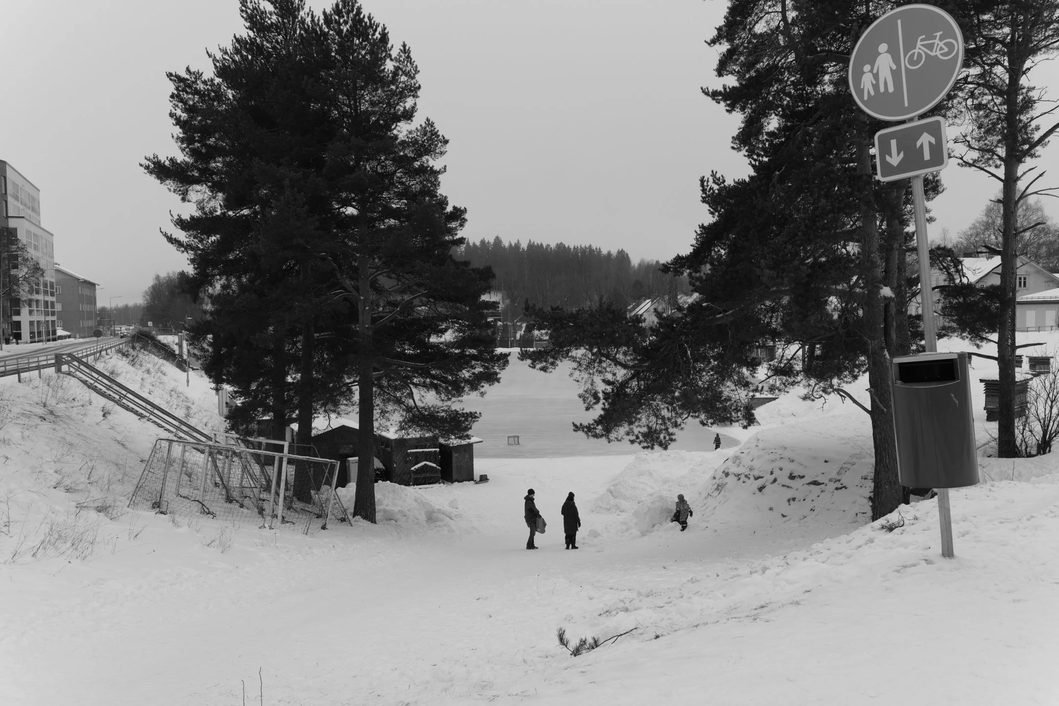 Luminen puistoalue, jossa on kolme ihmistä kävelemässä talvella. Taustalla on pieni jäälakeus ja puita, sekä rakennuksia. Oikealla on liikennemerkkilautakko ja roskakori.