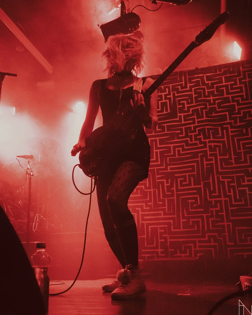 Nainen soittaa bassoa lavalla, taustalla geometrinen seinä ja lavavalot, hämärässä ja punaisessa valaistuksessa. Stoned Statues Lutakko.