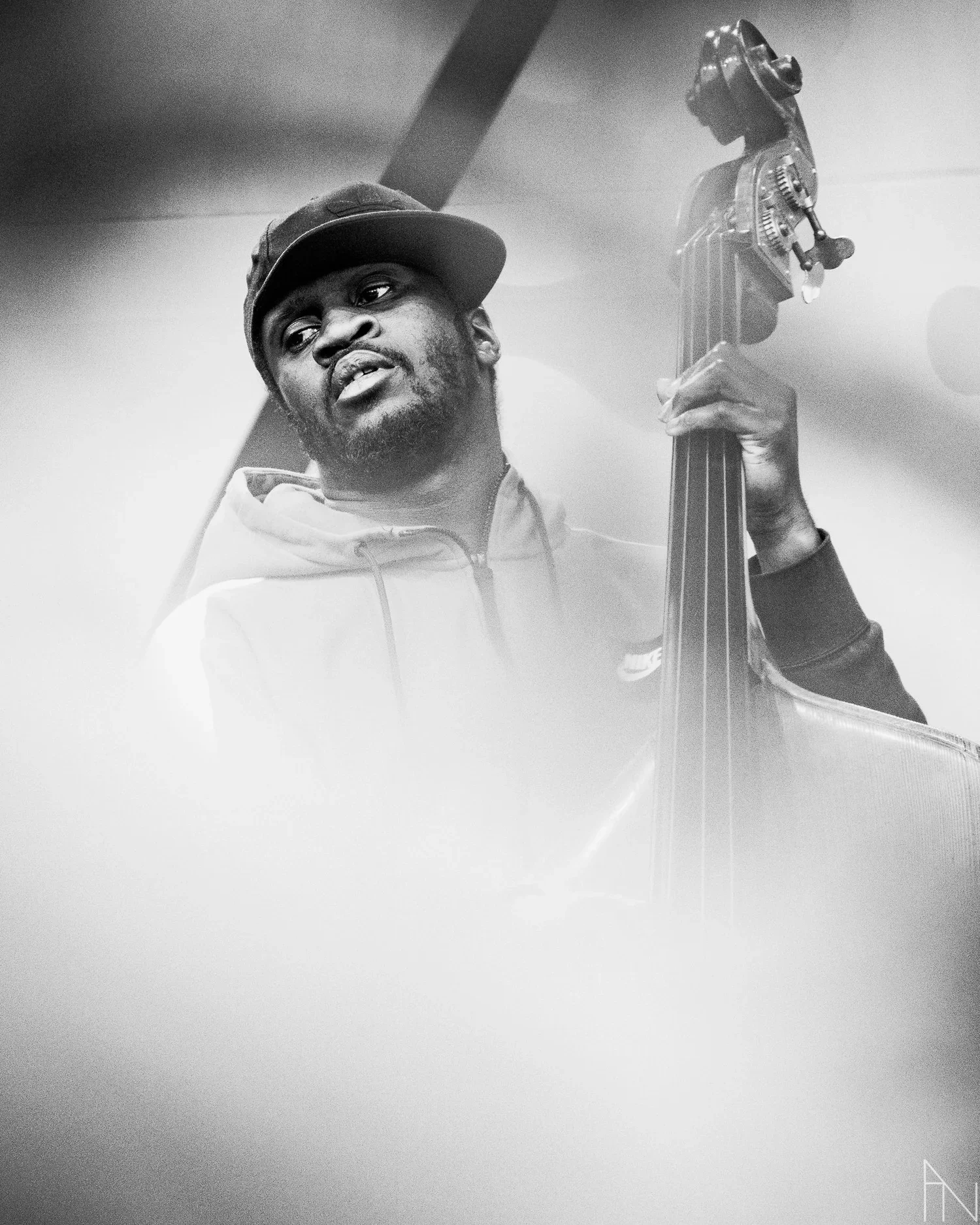 Mustavalkokuva miehestä, joka soittaa kontrabassoa, hän käyttää lippistä ja hupparia.