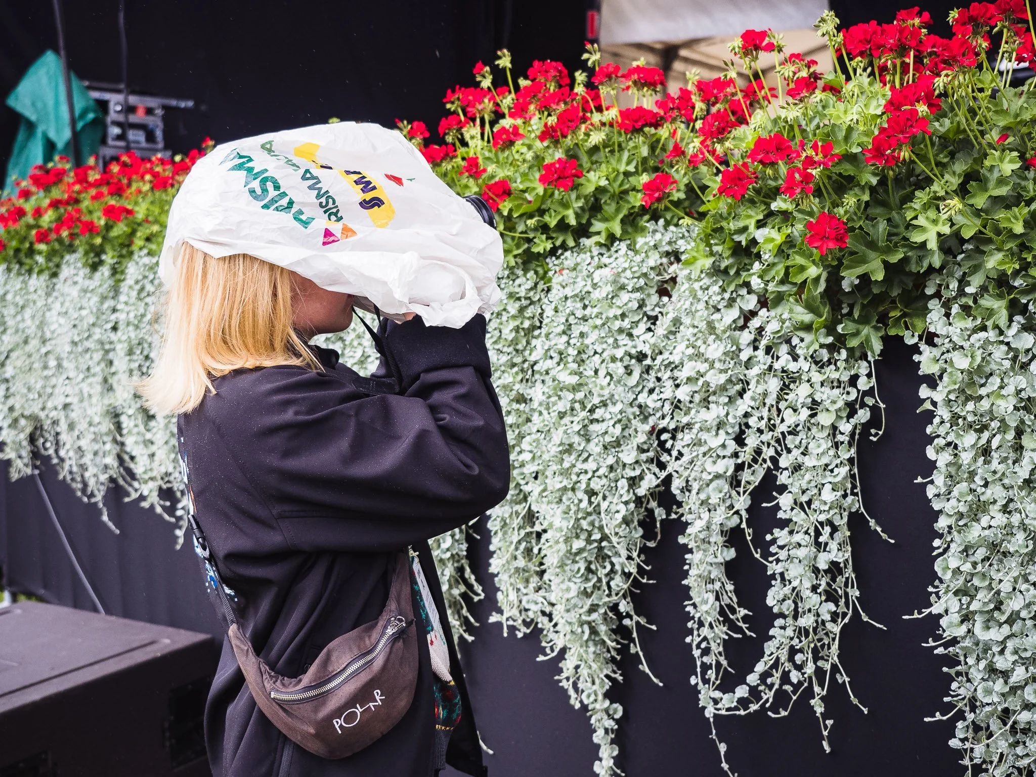 Nainen katsoo kukkivien kasvien päälle, käyttää suojaavaa päähinettä ja käsineitä, ja hänellä on pussi olalla.