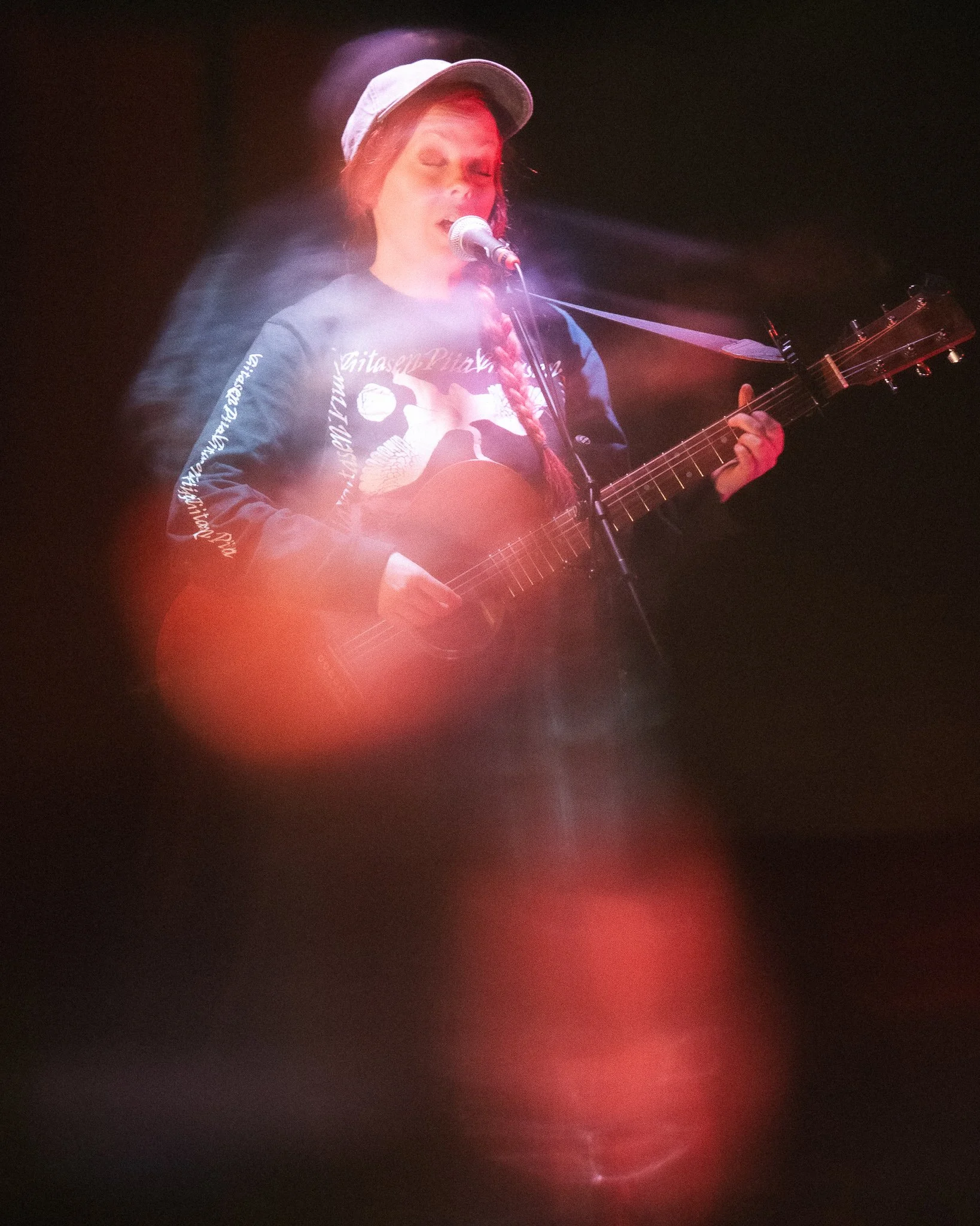 Nainen laulamassa kitara soittaen, pysähdyspaikassa pimeässä. Hänellä on farkkutakki ja lippis.