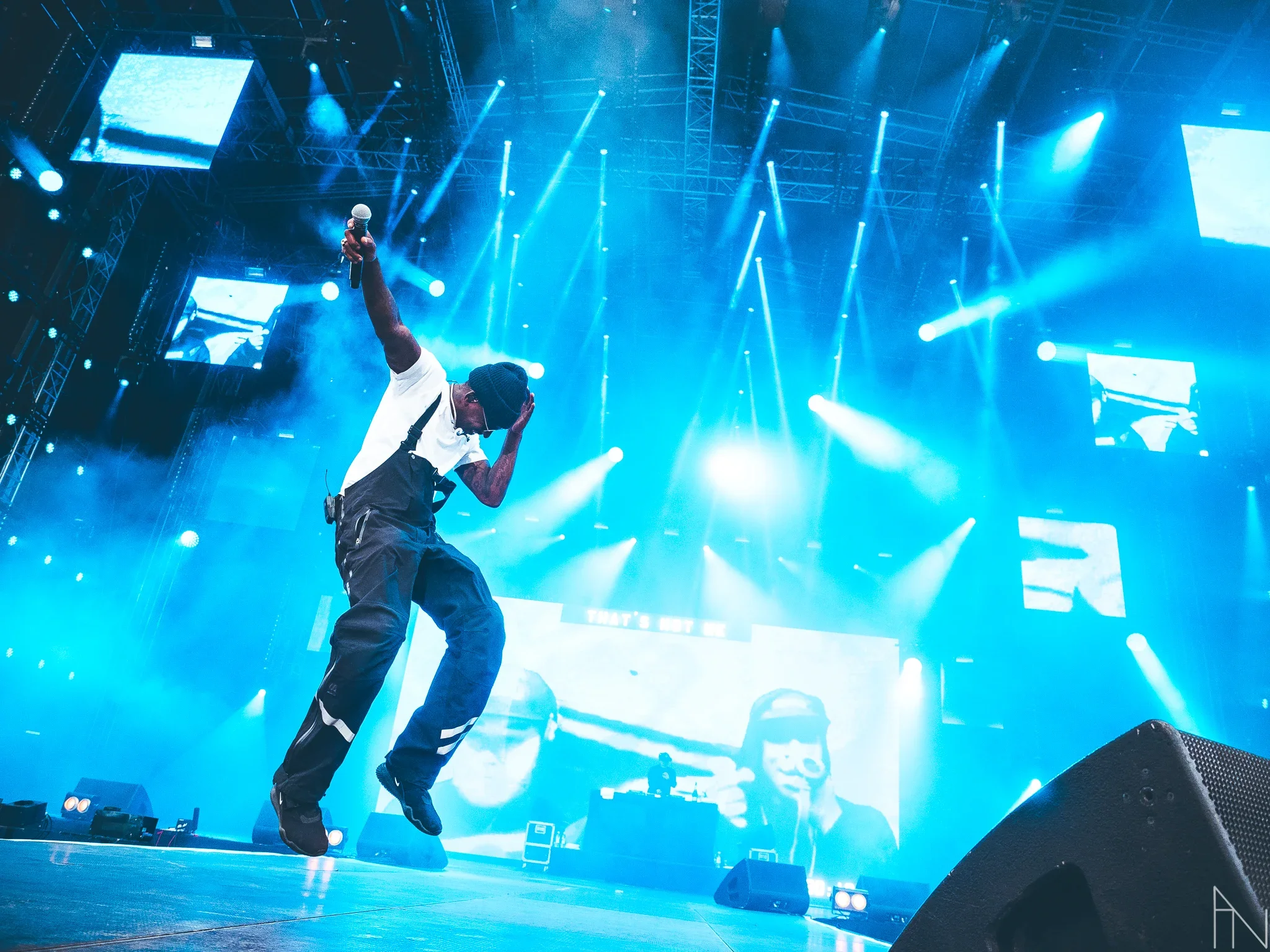 Mies esiintyy lavalla heittäen mikkiä ilmaan, taustalla suuret näytöt ja valotaustat, näkyy myös DJ ja kaksi muutakin ihmistä suurilla näytöillä.