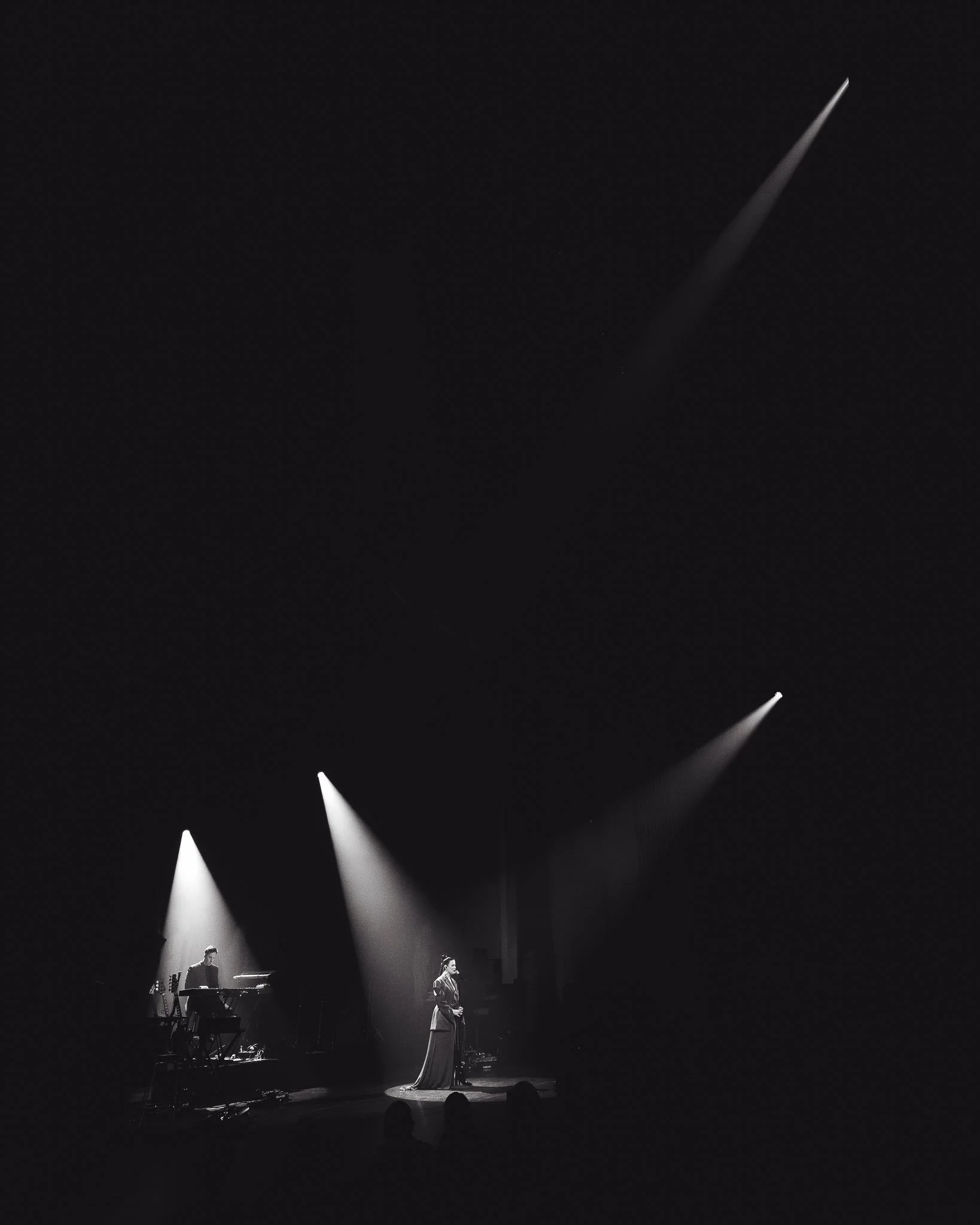 Musta ja valkoinen kuva teatteri- tai konserttisalin lavasta, jossa esiintyy nainen ja mies. Nainen seisoo keskellä ja mies soittaa koskettimia. Läheisiä valoja suuntautuvat kohti esiintyjiä.