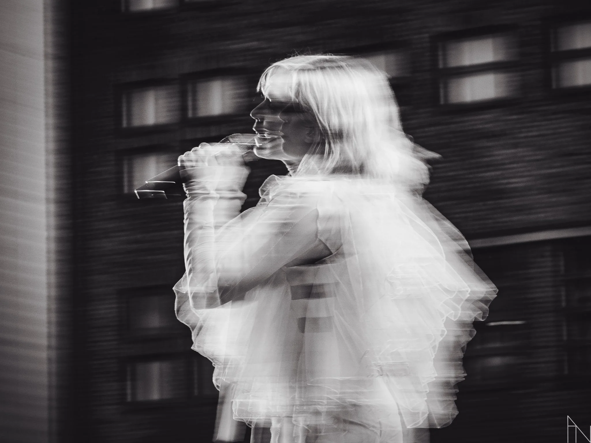 Mustavalkoinen kuva naisesta juomassa lasillista juomaa, taustalla rakennus. Kuvassa näkyy liikkeen sumentama vaikutus. Vesala YleXPOP 2022.