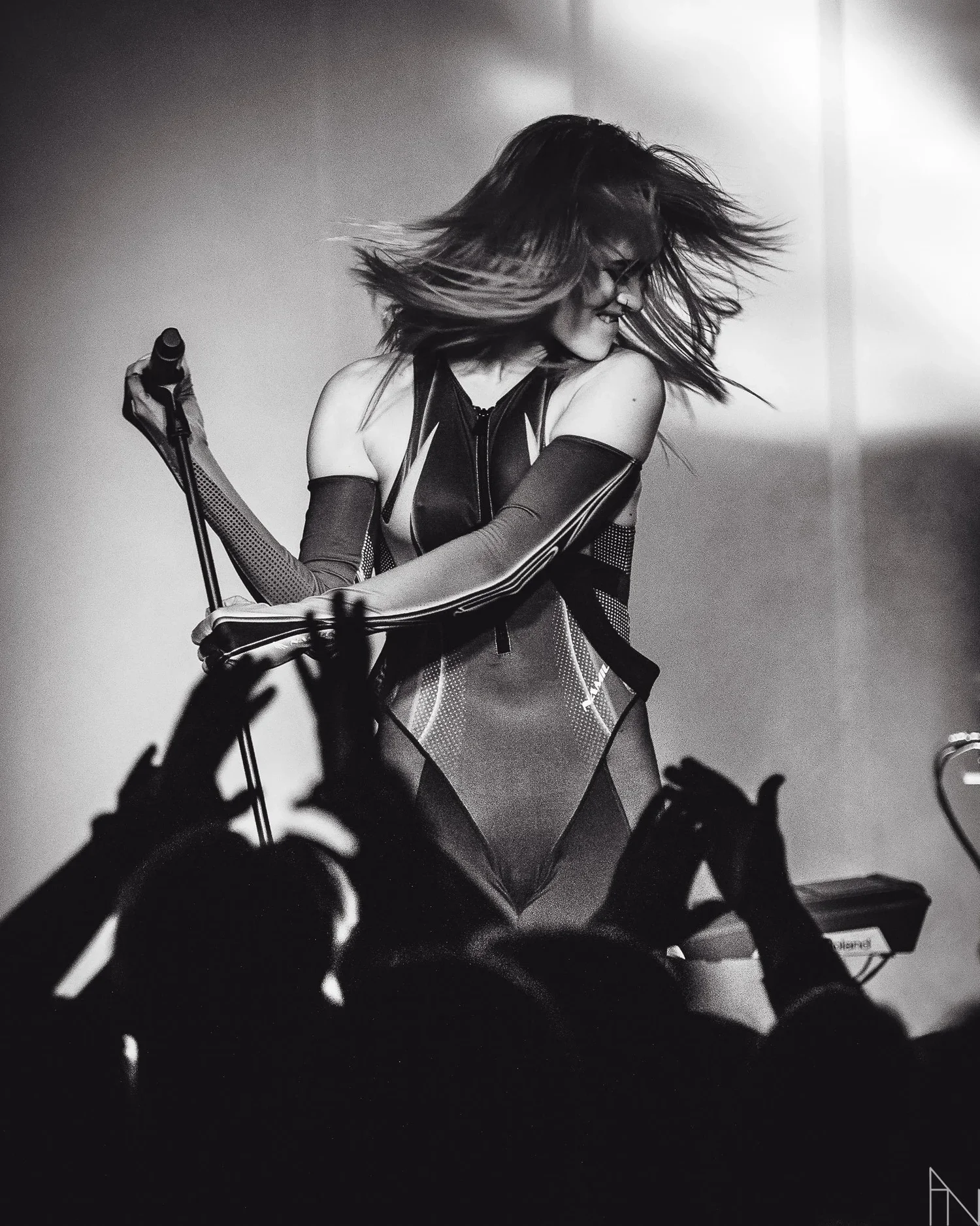 Mustavalkoinen kuva esiintyjästä lavalla, jossa on pitkät hiukset ja assymetrinen asu, ote mikrofonista, taustalla kirkas valo, yleisöä edessä.