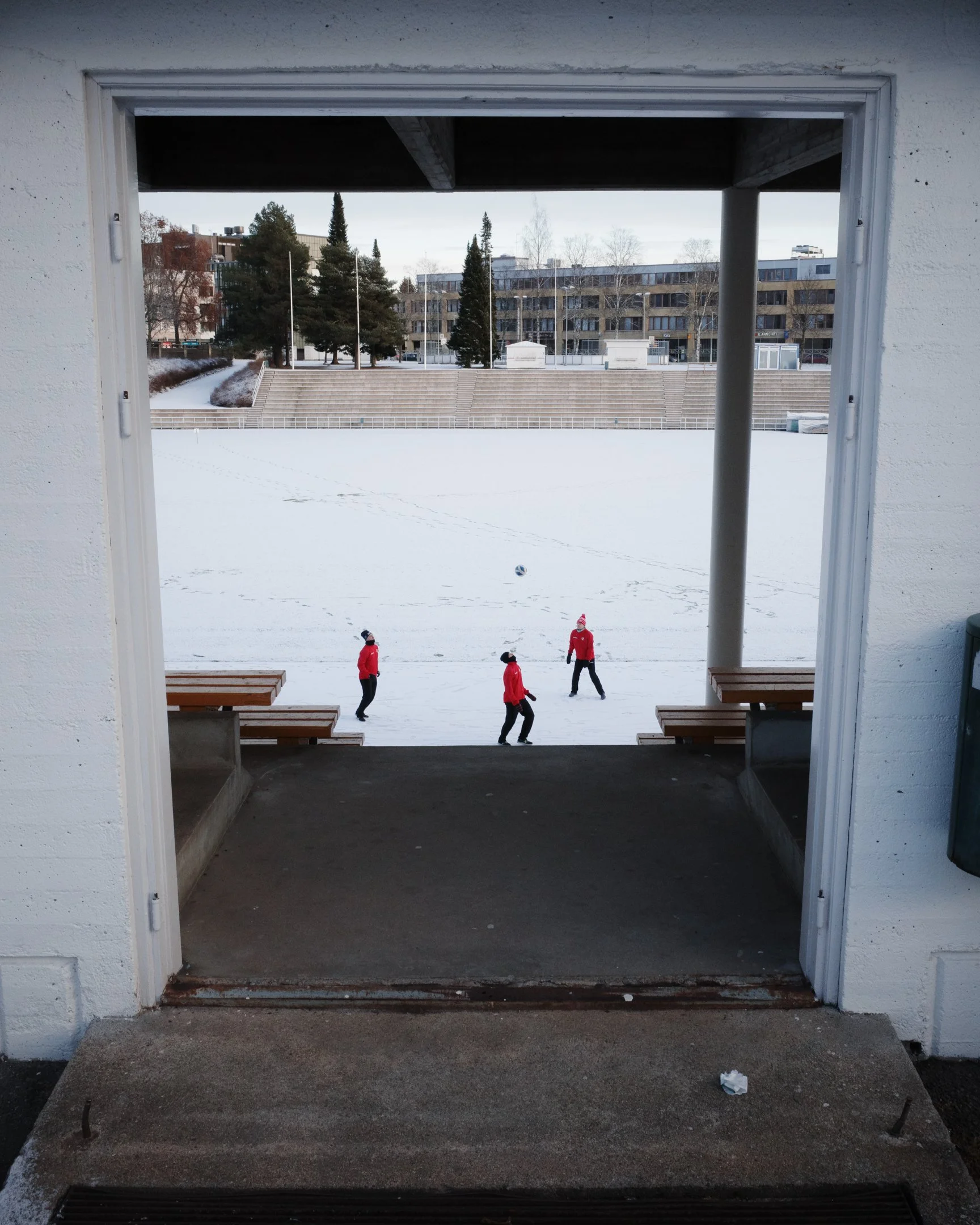 Kuvassa näkyy luminen urheilukenttä, jossa kolme henkilöä pukujen ja pipojen kanssa ovat pelaamassa jalkapalloa. Kuvan katseluikkuna rajaa näkymän sisään, ja näkymä sisältää portin, jonka edessä on betonilattia ja penkkejä.