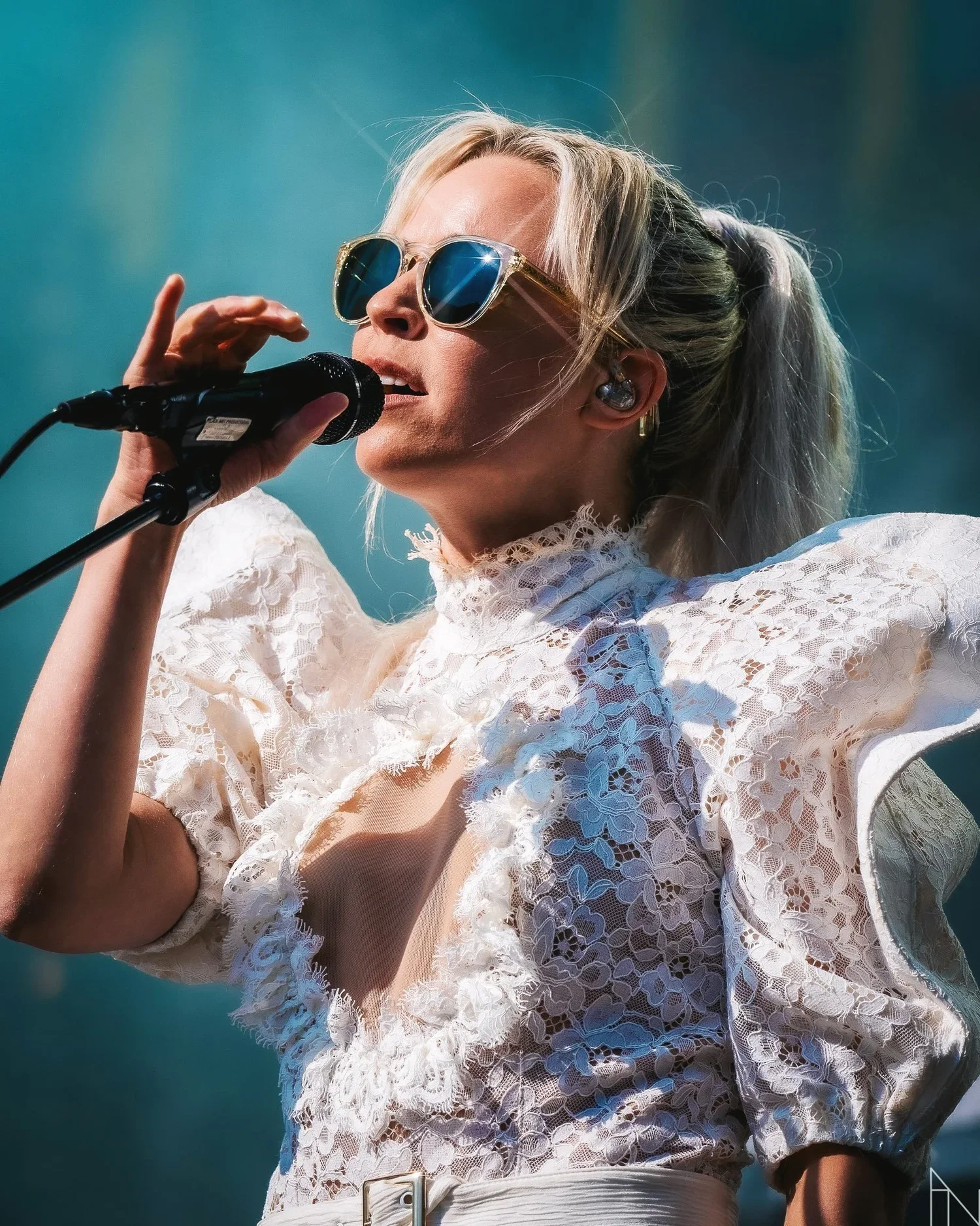 Nainen laulaa mikrofonin kanssa konserttia, käyttää aurinkolaseja ja on lippalakkimainen puku päällään.