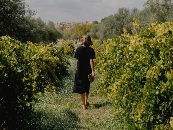 A woman is sat in a wooden chair, writing in a journal placed on her lap, with a cup of black coffee on a wooden table next to her.