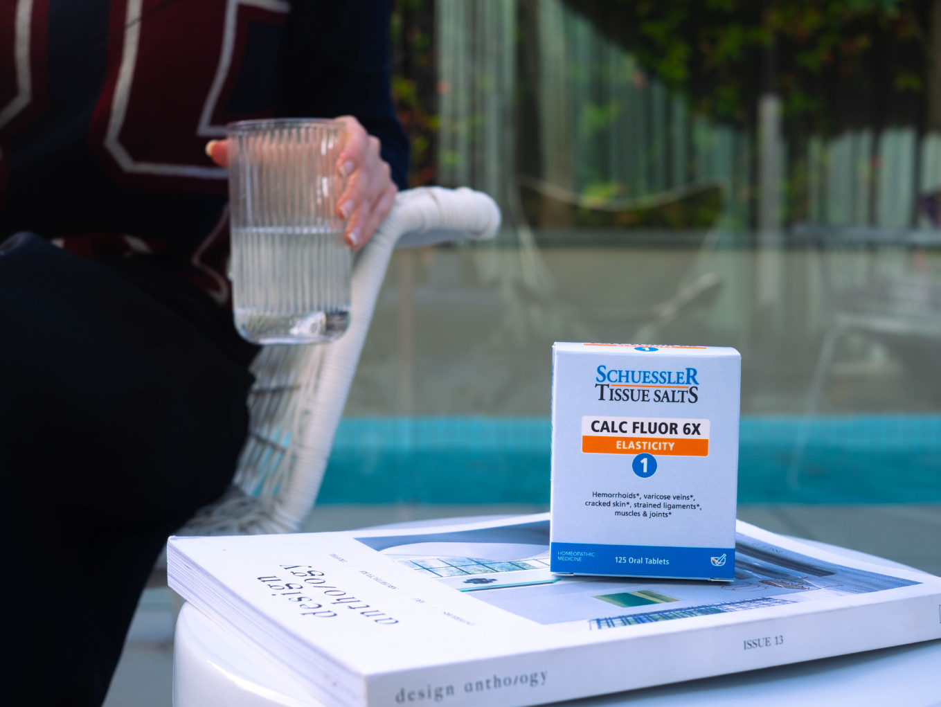 A box of Tissue Salts placed on top of a design magazine, with a woman in a black shirt holding a glass of water in the background.