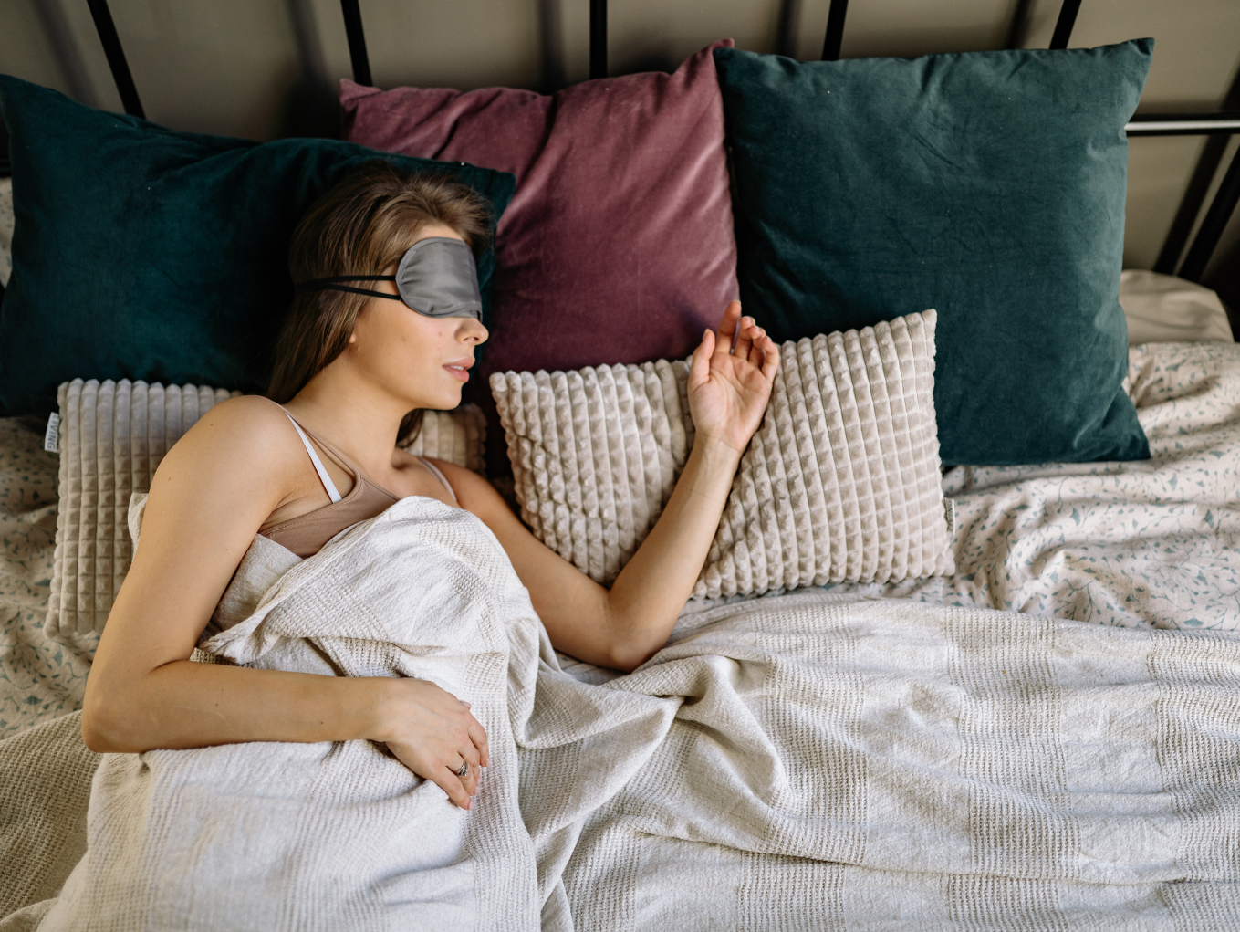 A woman asleep in bed with her head resting on a green and a red pillow. The woman is wearing a grey eye mask.