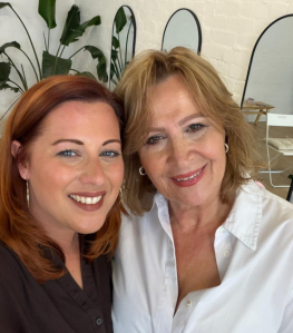 Two women smiling for a selfie indoors, with plants and mirrors in the background.