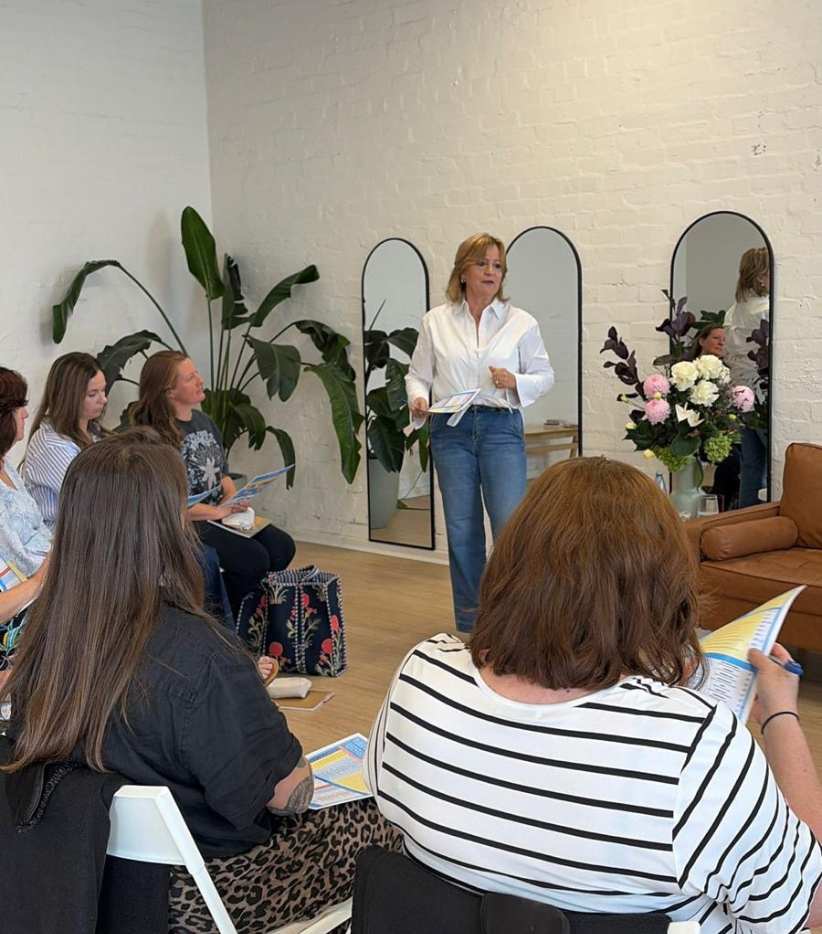 A woman standing and speaking to a group of women seated in a room with white brick walls, large mirrors, and green plants, during a presentation or class.