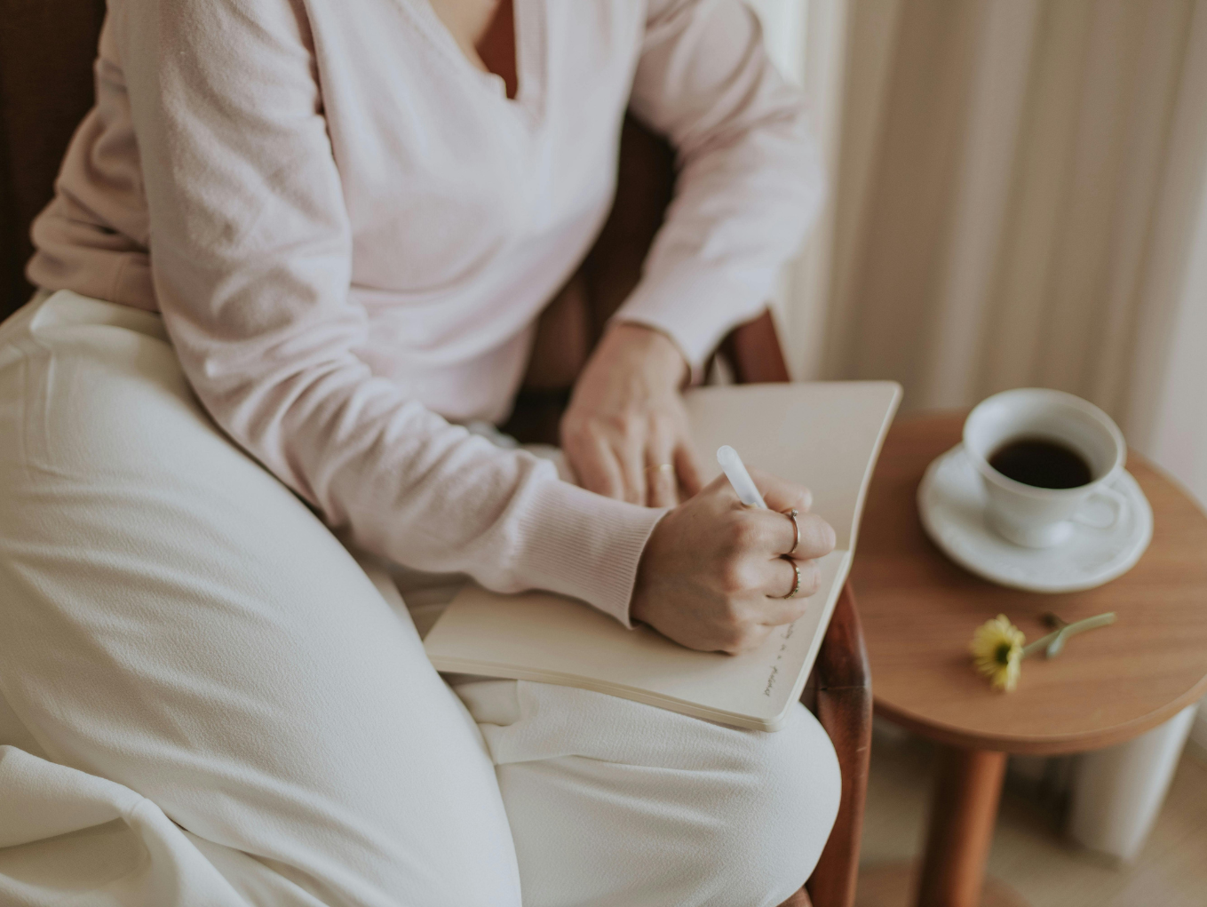 A woman is sat in a wooden chair, writing in a journal placed on her lap, with a cup of black coffee on a wooden table next to her.