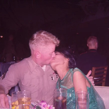 A man in a military uniform and a woman are kissing at a table with drinks in a dimly lit setting.
