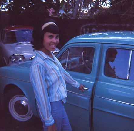 A woman with dark hair, wearing a striped shirt, standing next to a vintage blue car.