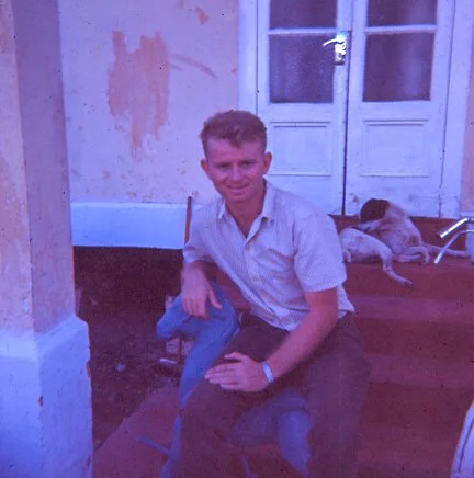 A young man sitting outdoors on a step with a dog lying beside him, in front of a white door and a wall with peeling paint.