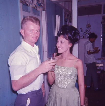 A man and woman at a gathering, holding drinks and smiling at each other, with another person in the background.