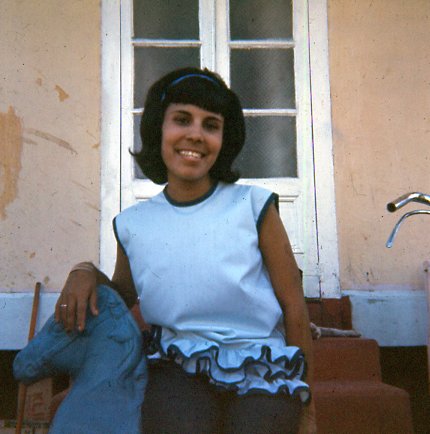Young girl with short black hair smiling, sitting outdoors near a window with a light-colored wall in the background.