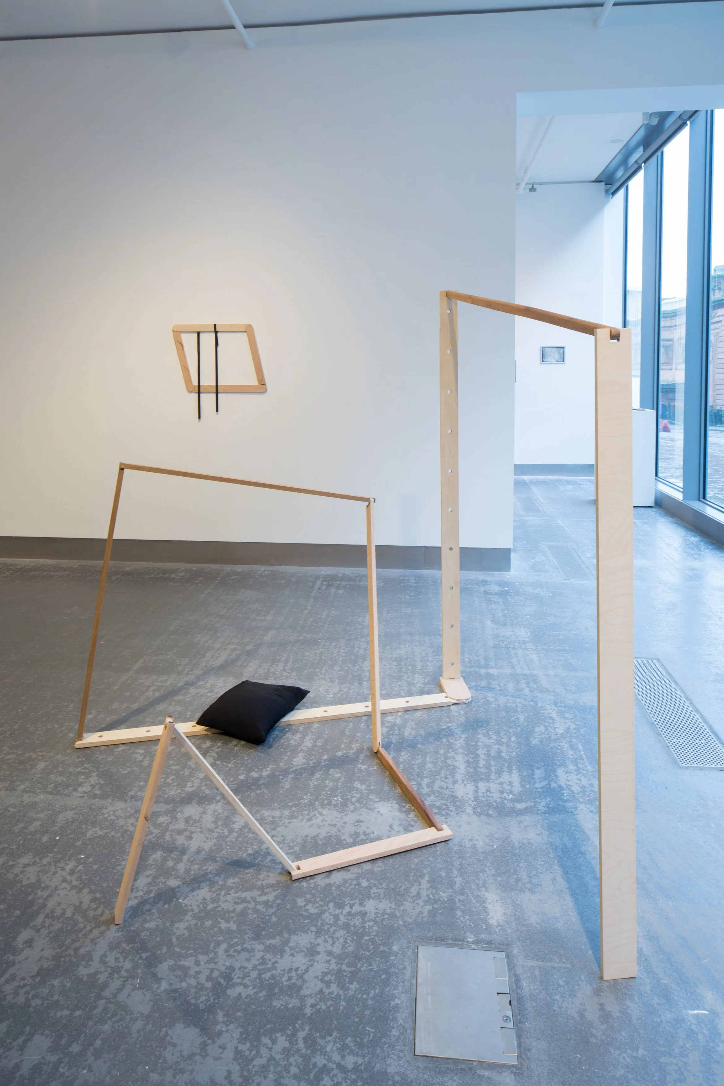 Interior view of an art gallery or exhibition space with wooden frames and structures, and large floor-to-ceiling windows on the right side, letting in natural light.
