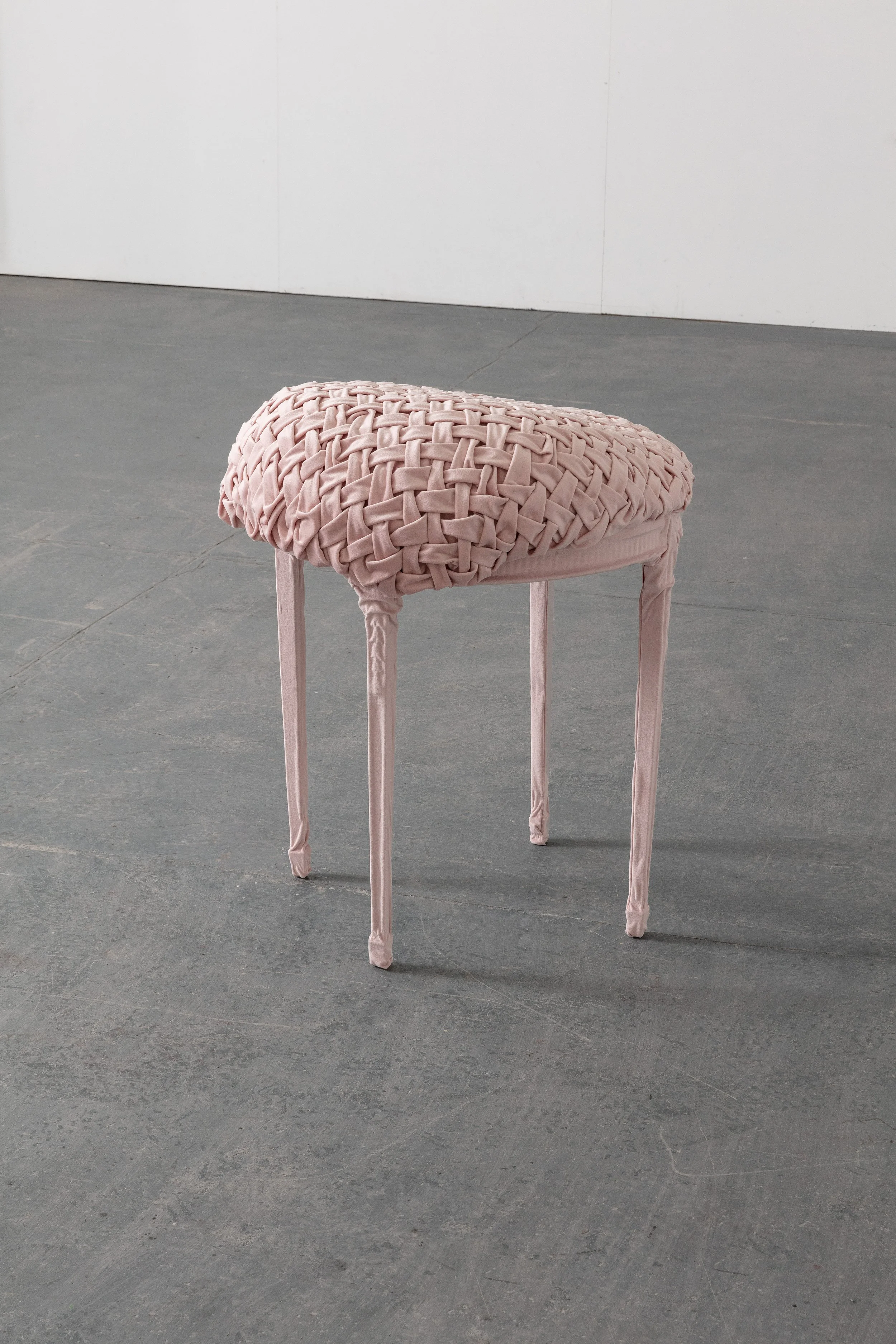 Pink woven fabric upholstered stool with four slender legs, in an empty room with gray concrete floor and plain white walls.