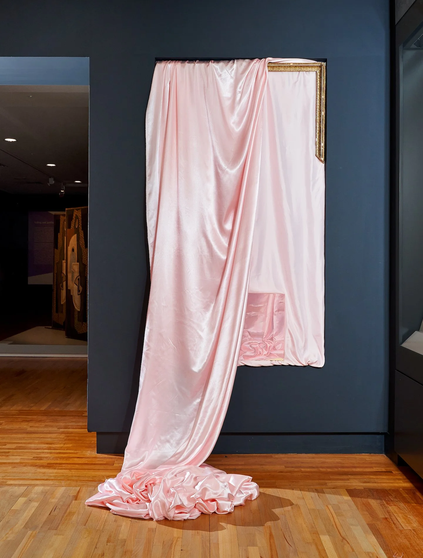 A framed artwork covered with pink satin fabric that drapes down and pools on the wooden floor.
