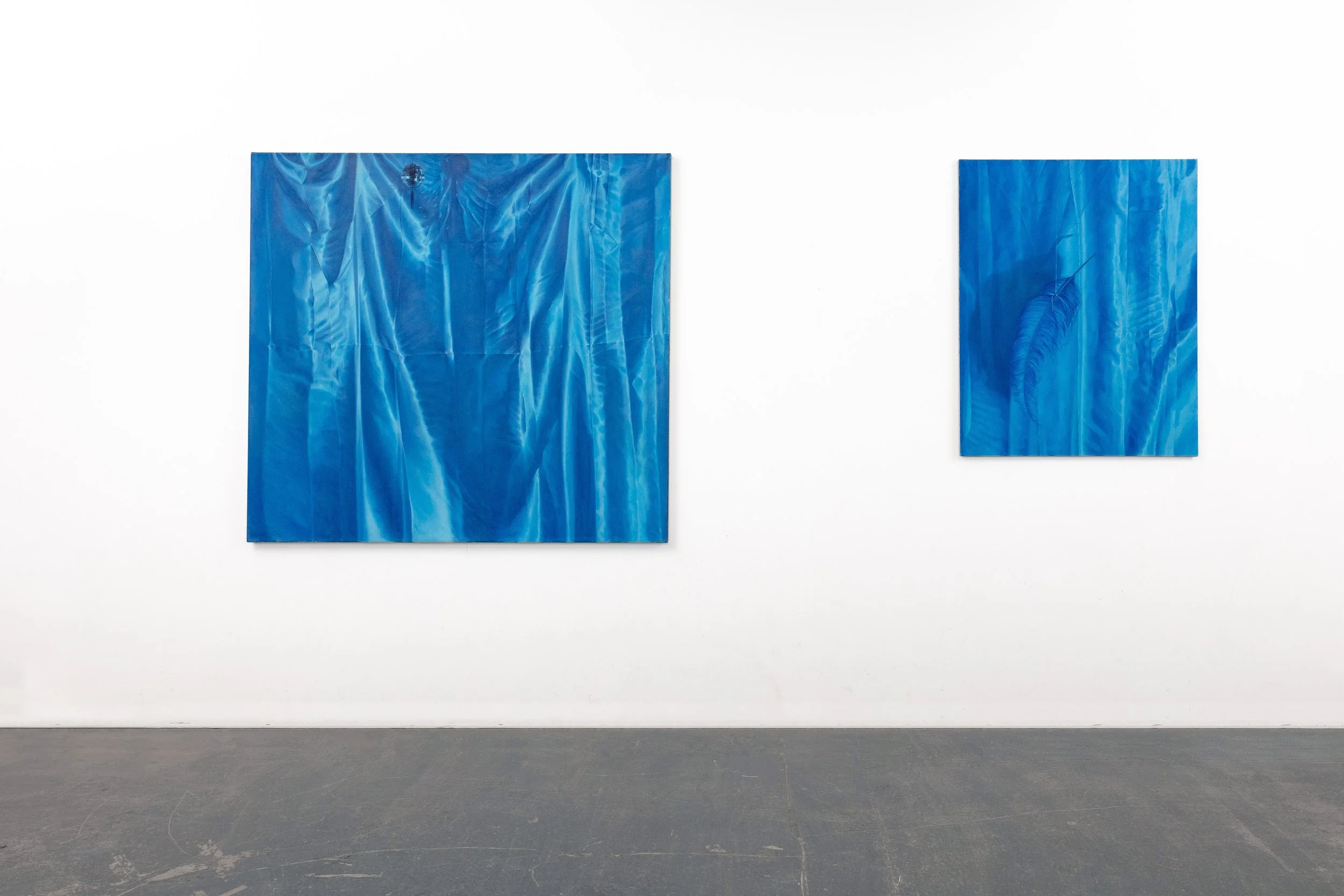 Two blue abstract paintings on a white gallery wall, with a dark gray floor below.