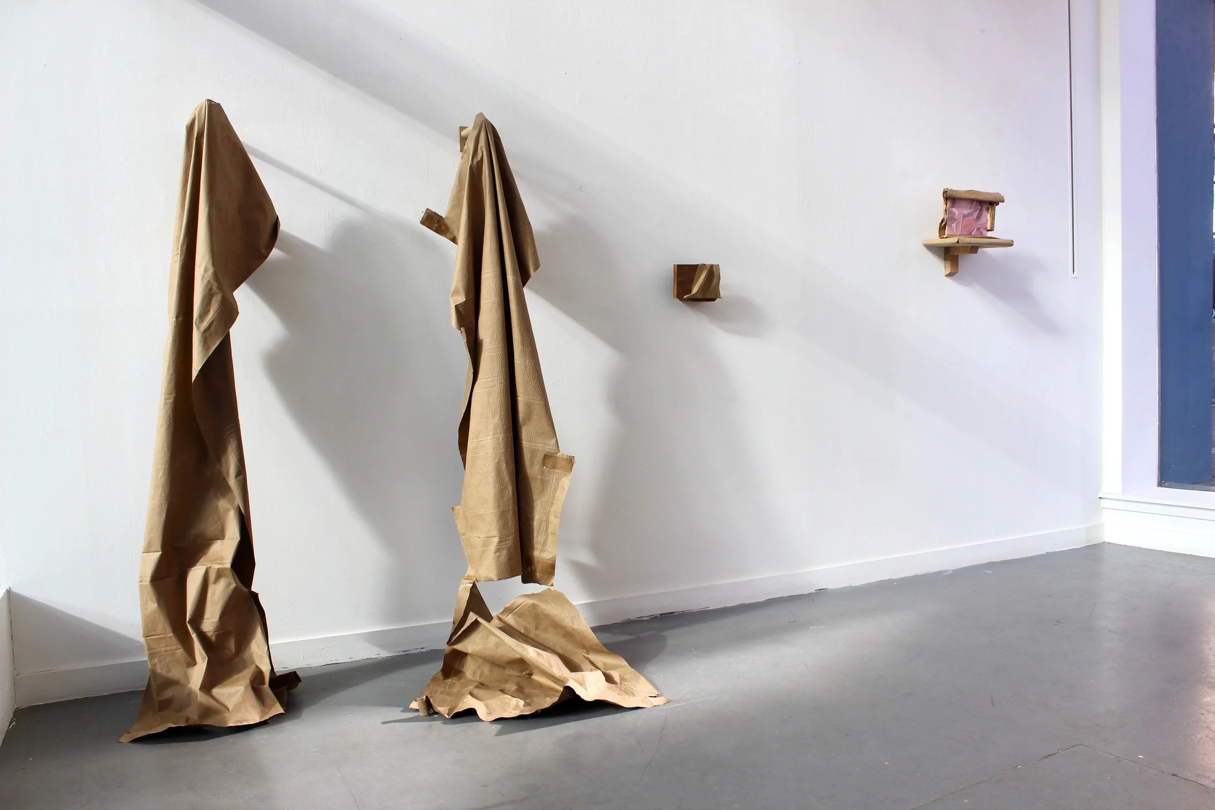 Paper bags and brown paper wrapping on the floor and hanging on the wall, with a small shelf holding a pink object in a wrapped package, set against a white wall and gray floor.