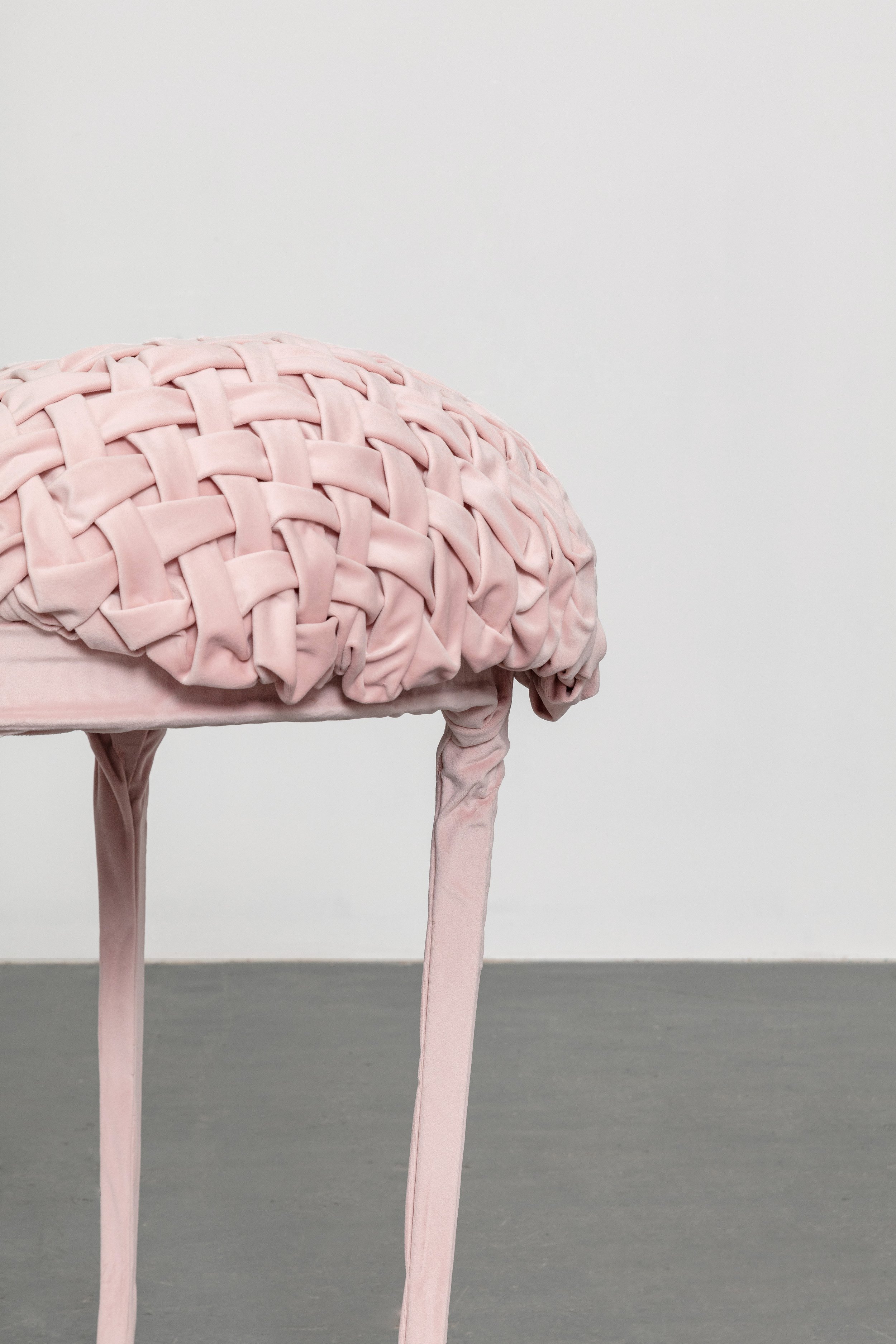 Sculpture resembling a brain, made from pink fabric with a woven pattern, standing on four pink fabric-covered legs, against a plain gray and white background.