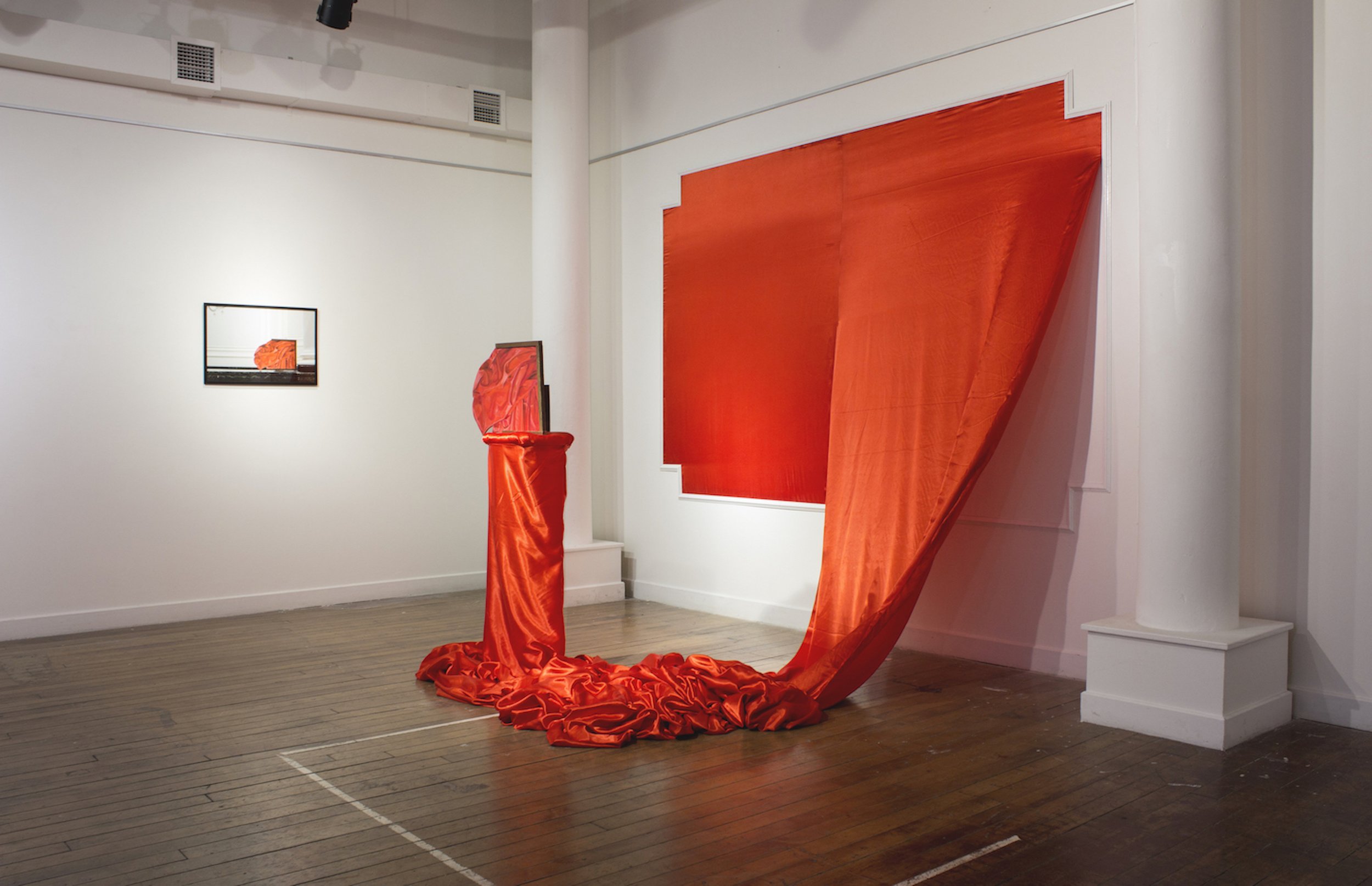 An art installation with large orange fabric draped over a frame and floor, with smaller orange fabric-covered objects on the floor in an art gallery or museum setting.