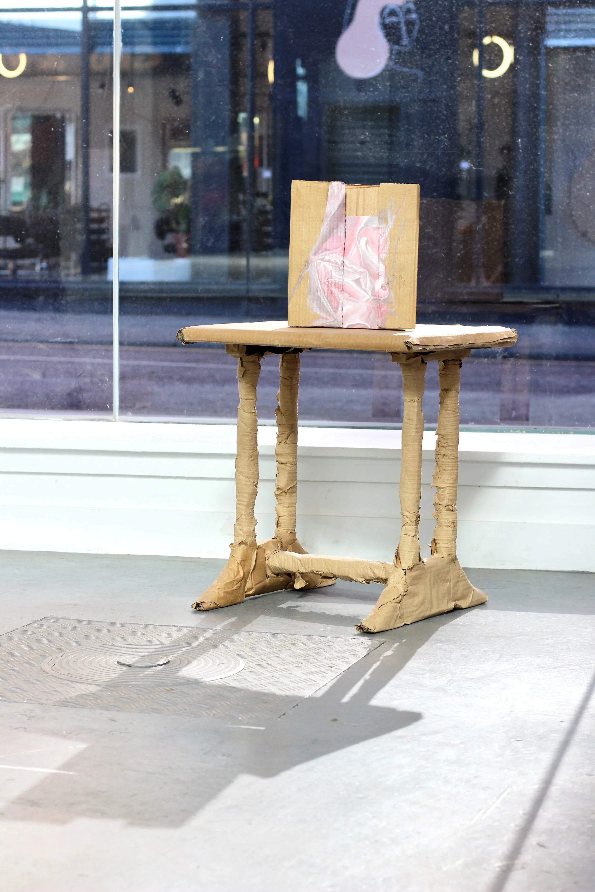 A cardboard table with cardboard legs and a cardboard top, with a cardboard box sitting on it, located in front of a large window.
