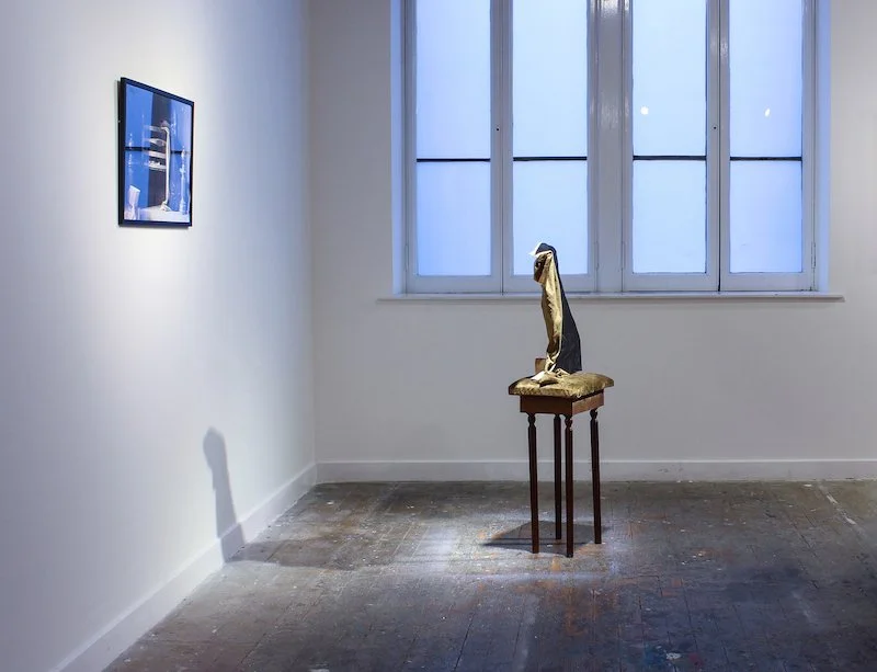An art gallery with a painting of black cloth on a small wooden table, and a framed photo on the white wall to the left.