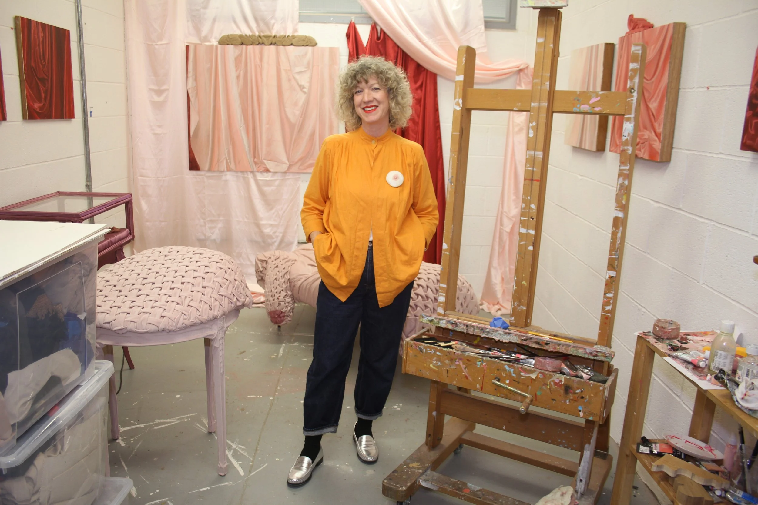 A woman with curly blonde hair wearing a yellow jacket and silver shoes standing in an art studio with paintings, canvases, and art supplies around her.