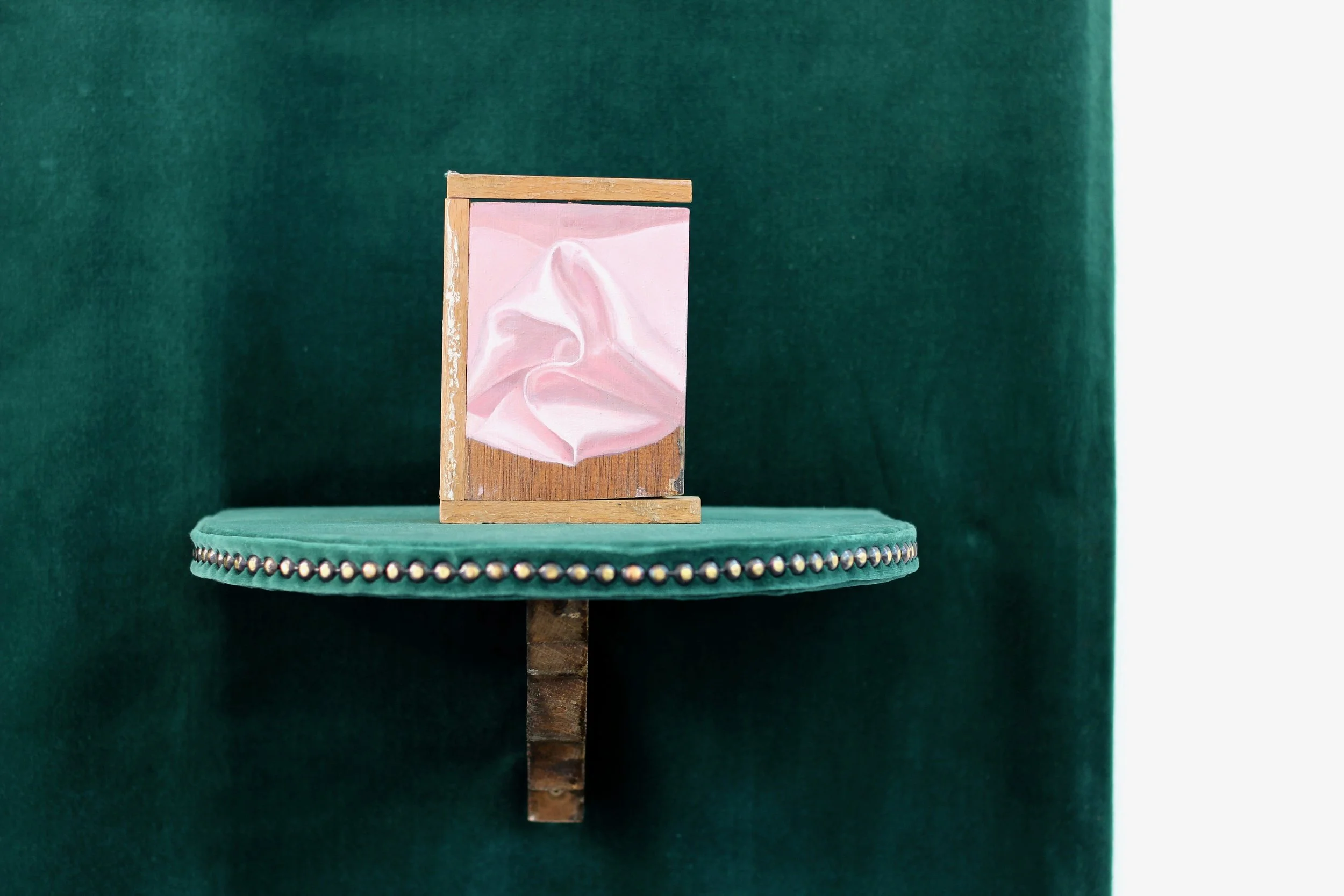 A small painting with a pink background and a white, abstract, flowing shape, displayed on a wooden easel. The easel is on a green, velvet-covered round table with decorative silver studs around the edge, set against a dark green fabric backdrop.