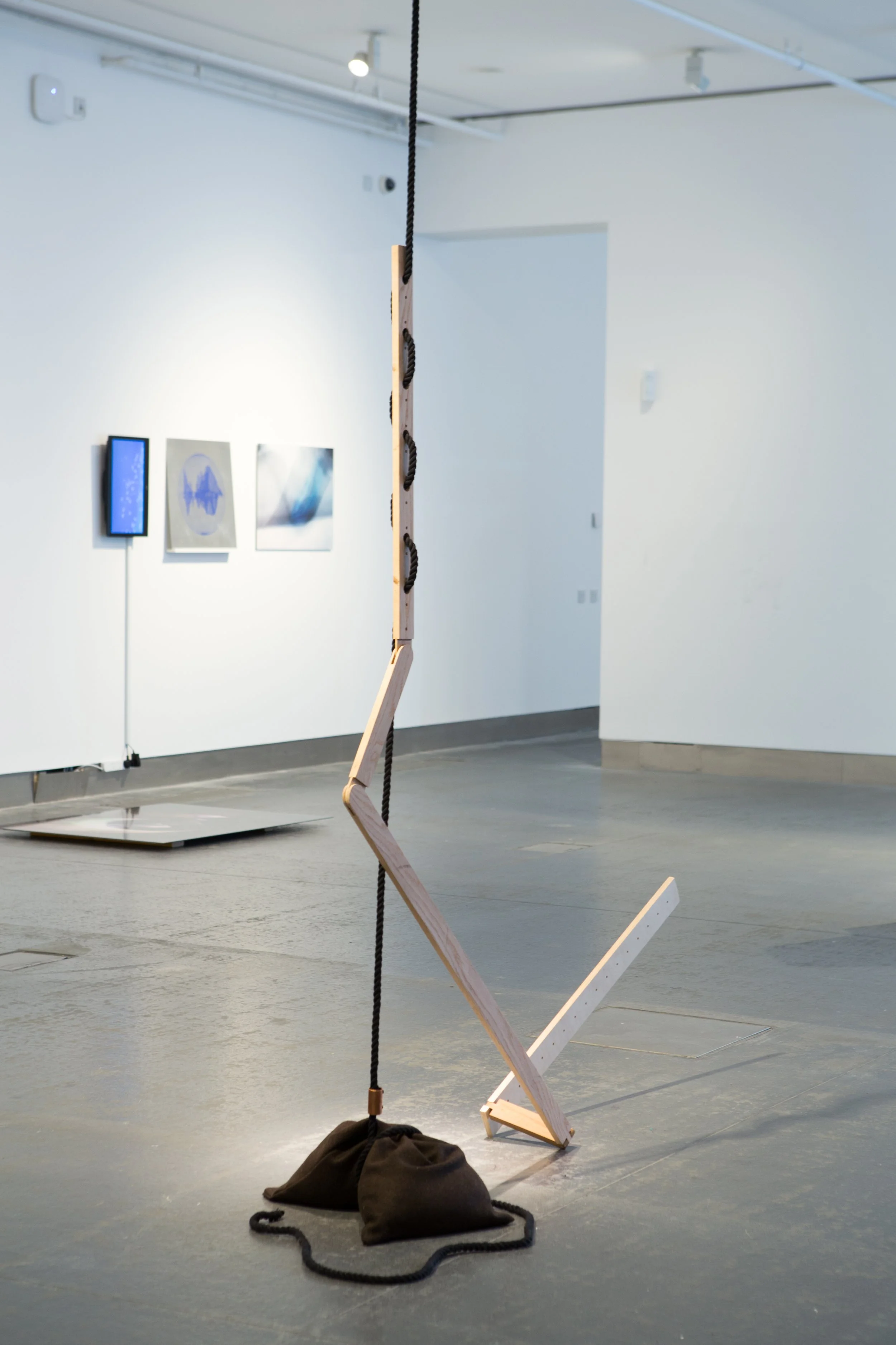 An art installation in a gallery featuring a wooden structure with black rope and a black bag on the floor with a rope attached. In the background, there are three abstract art pieces hanging on a white wall.