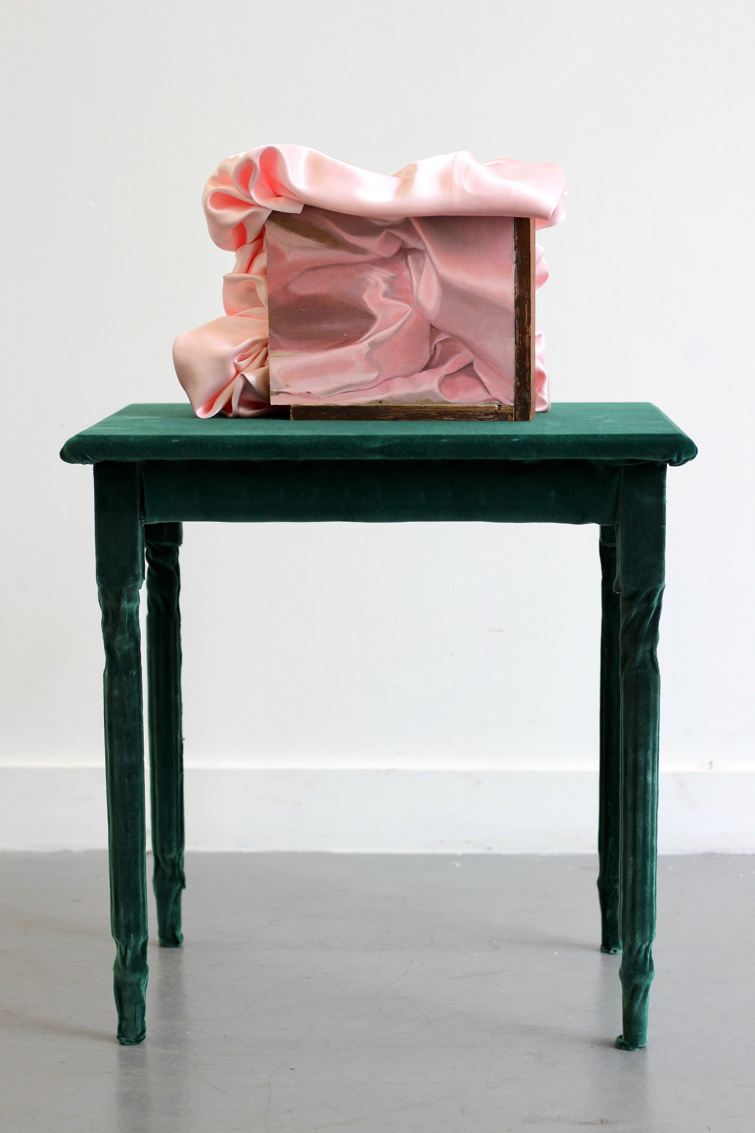 An art piece consisting of a pink satin-like fabric draped over a wooden box, placed on a dark green velvet-covered table against a plain white wall.
