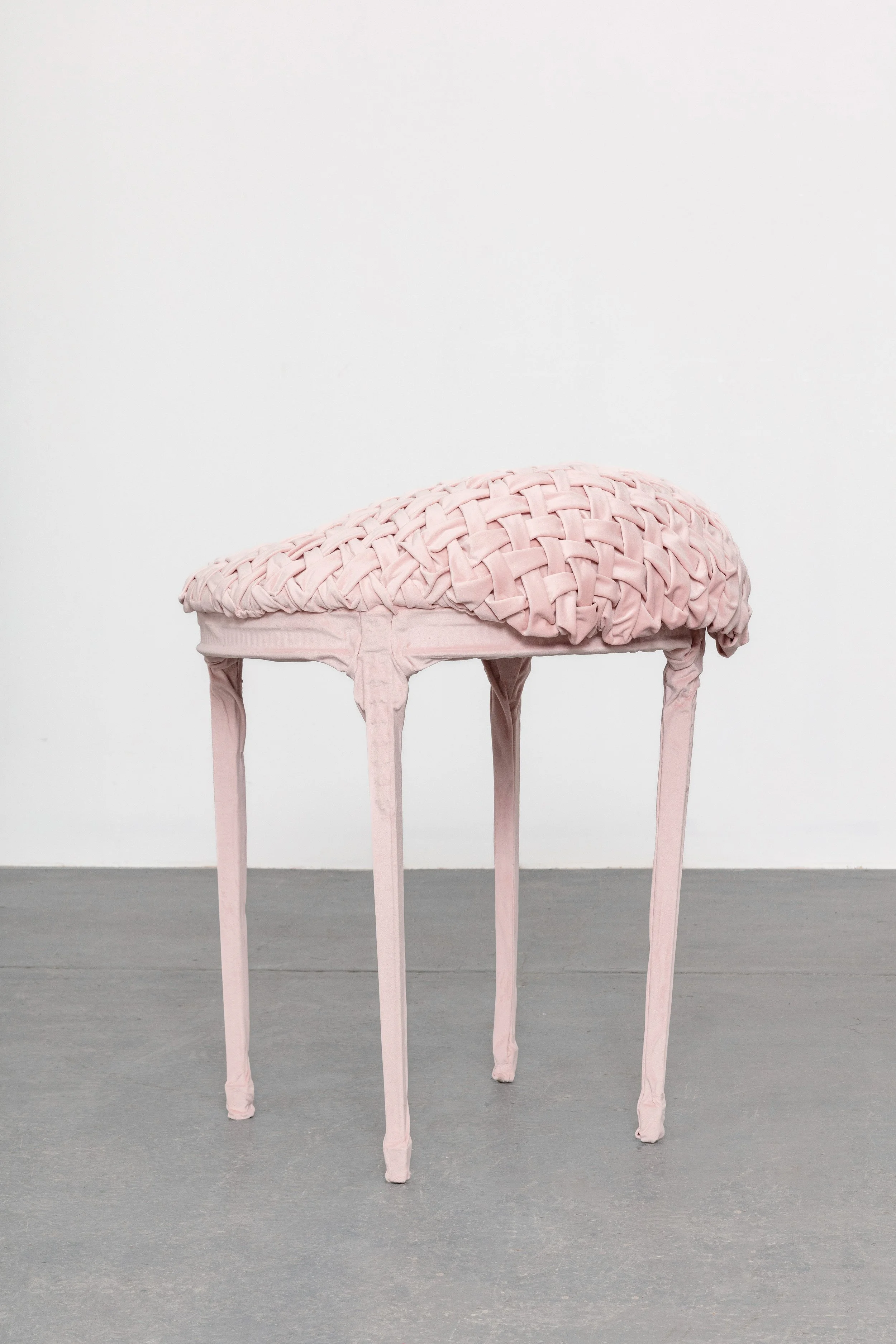 A pink, woven, cushion-like object resting on a tall, slender, pink wooden table with four legs, set against a plain white wall.