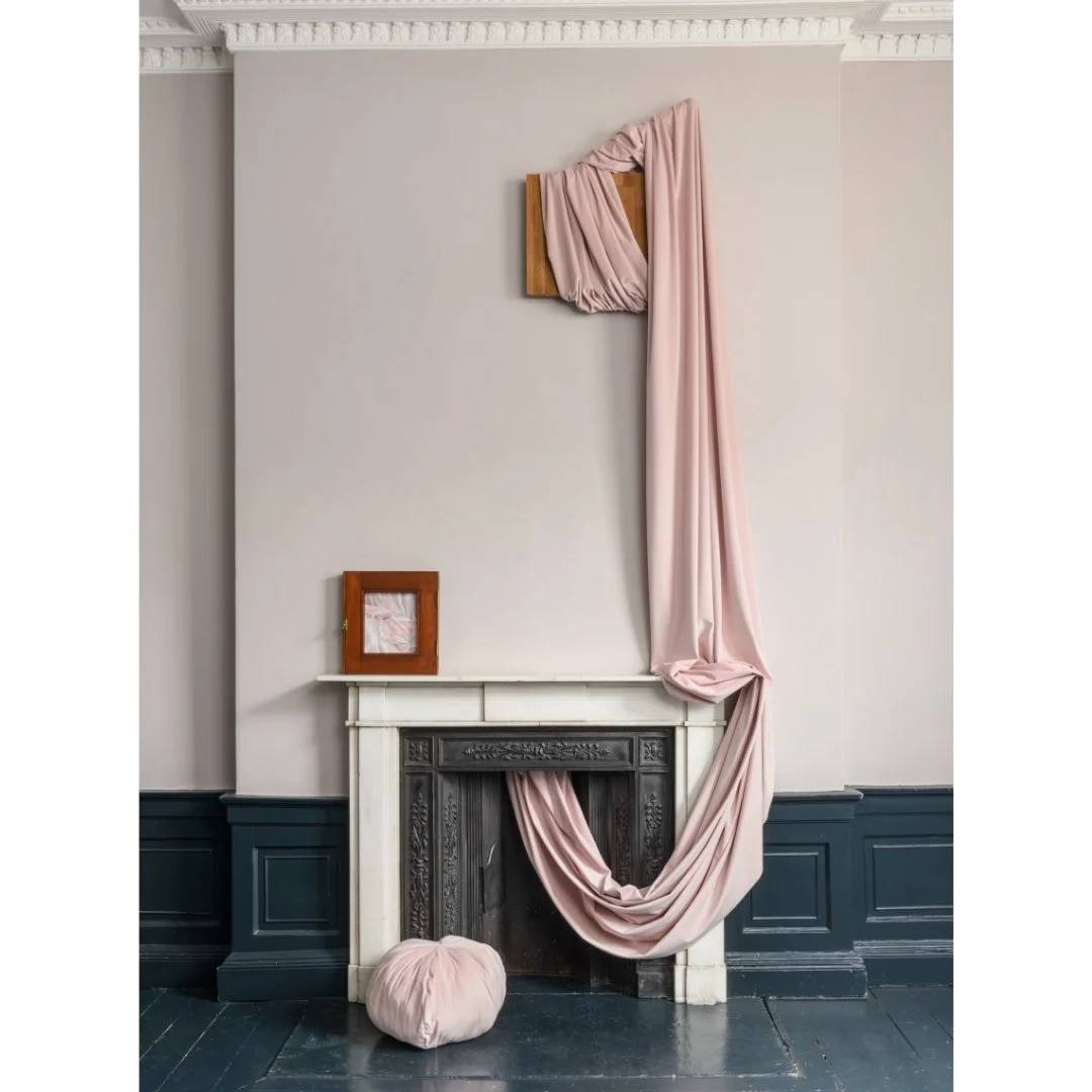 A decorative pink fabric draped over a wooden picture frame on a white wall, flowing down over a fireplace mantel and pooling onto the floor with a pink cushion in front.