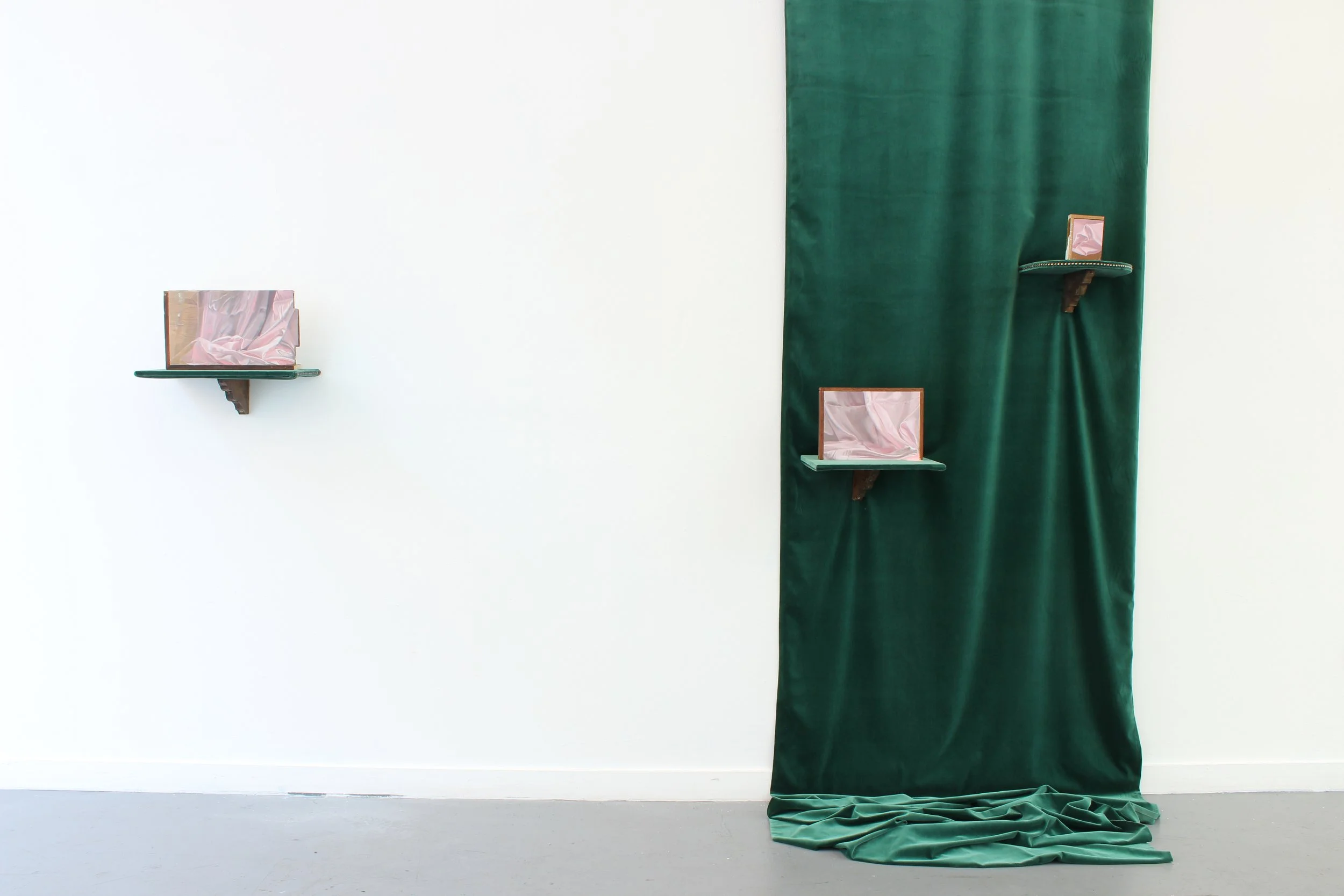 Cream-colored wall with three small shelves holding pink silk fabric-covered objects, green curtain on the right covering part of the wall, fabric pooling on the floor.