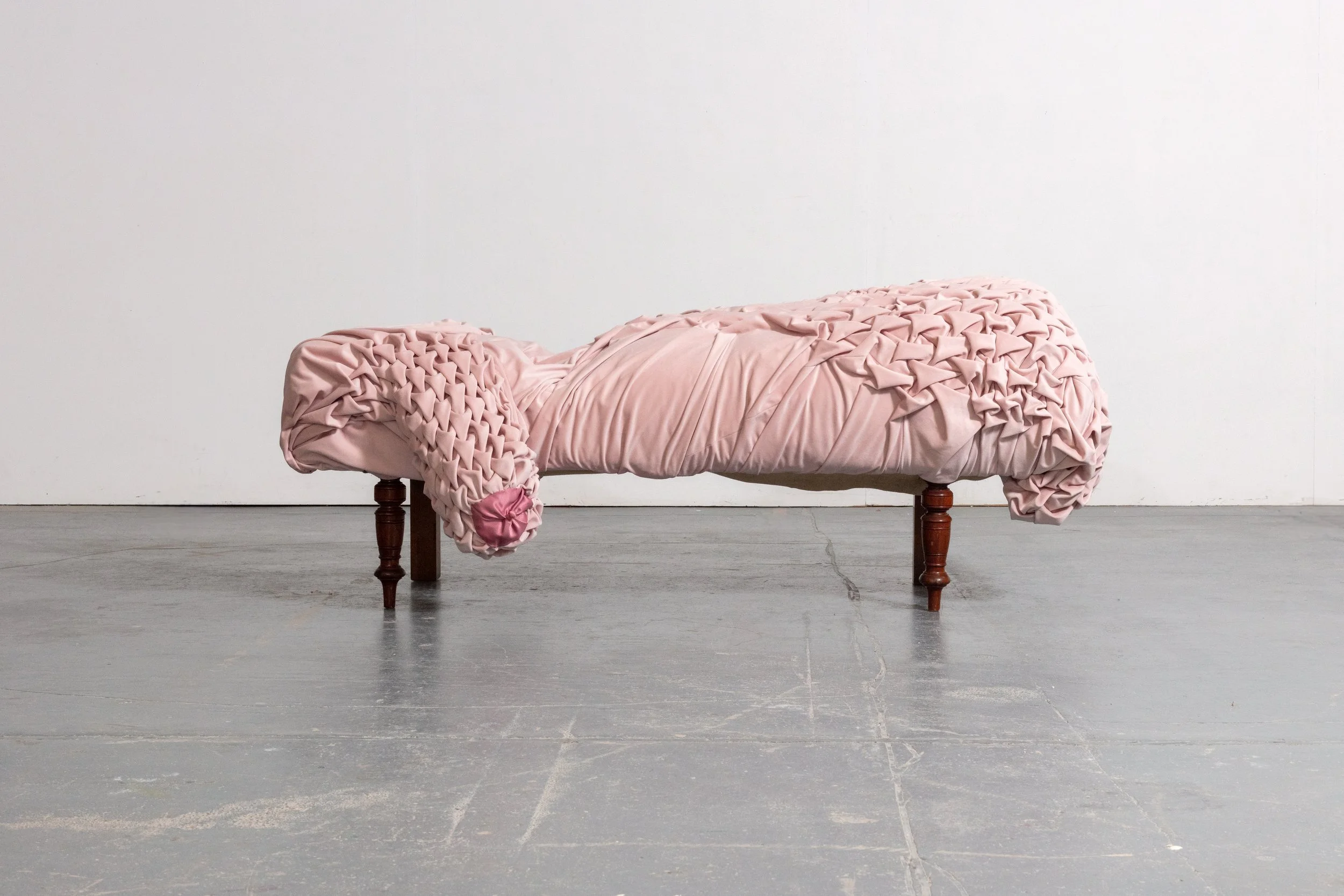 A pink, textured fabric sofa with wooden legs is positioned against a white wall and gray wooden floor.