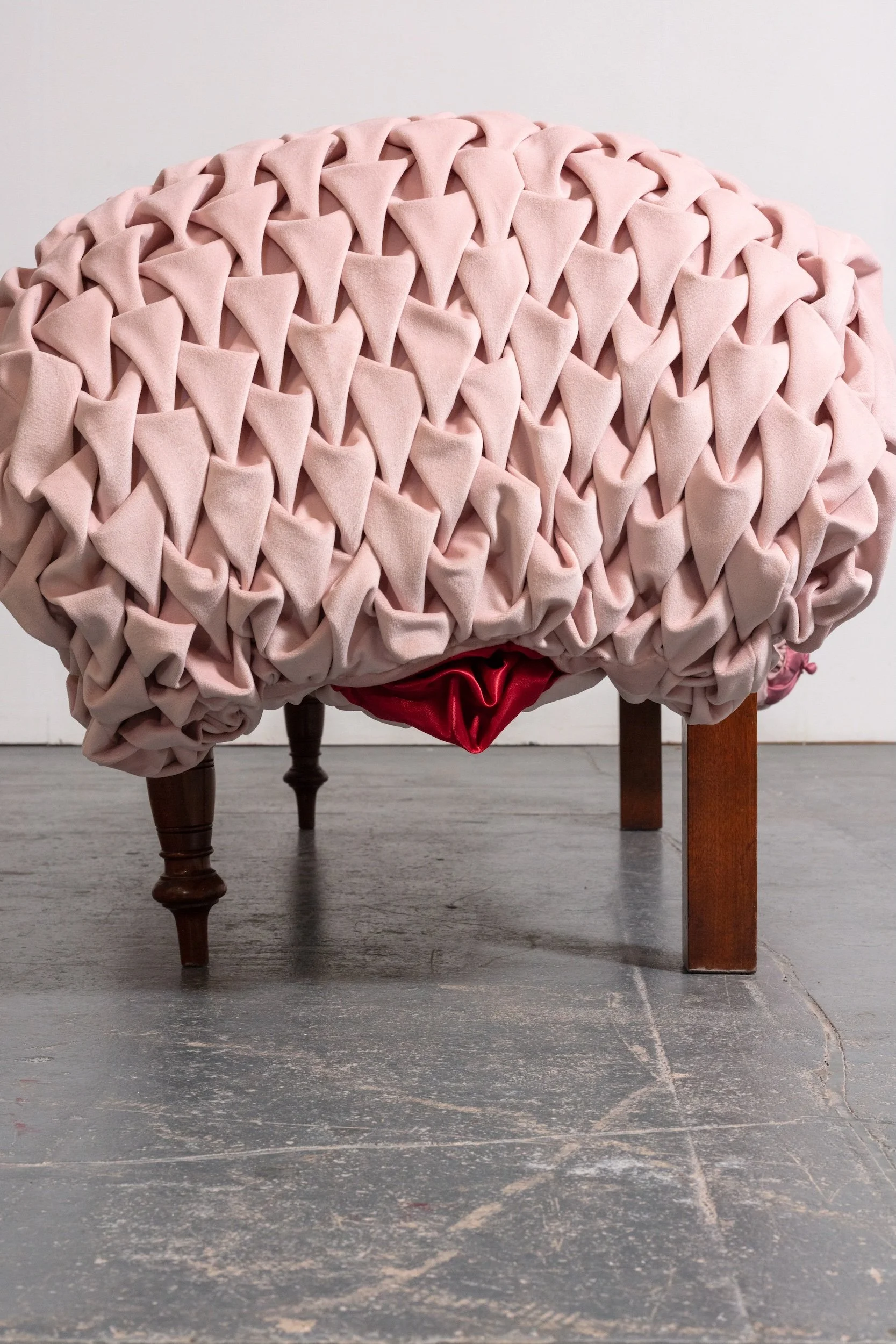 Close-up of a piece of furniture with a pale pink, quilted, tufted fabric covering, supported by wooden legs on a gray wooden floor.