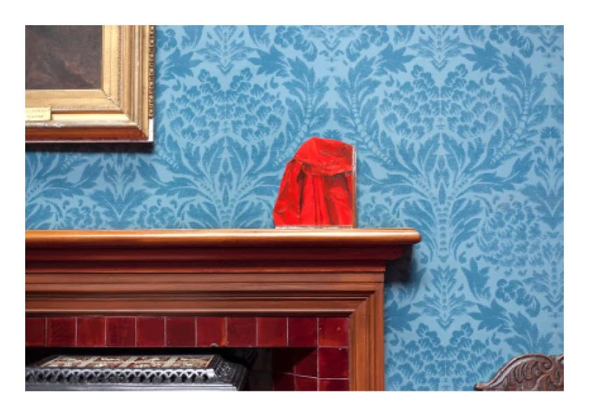 A red velvet gift bag sits on a wooden fireplace mantle in a room with blue damask wallpaper, a painting in a gold frame partially visible on the wall.