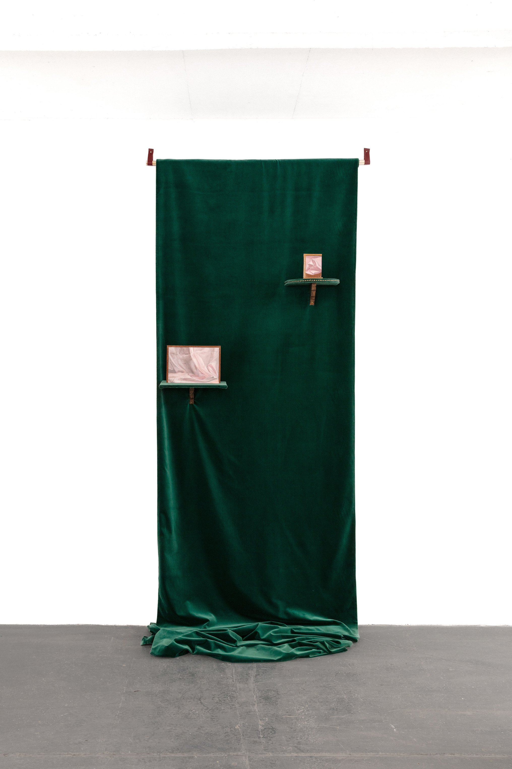 A dark green fabric backdrop hanging from a rod in an exhibition space with a gray concrete floor. There are two small shelves attached to the backdrop, each holding a pink object.
