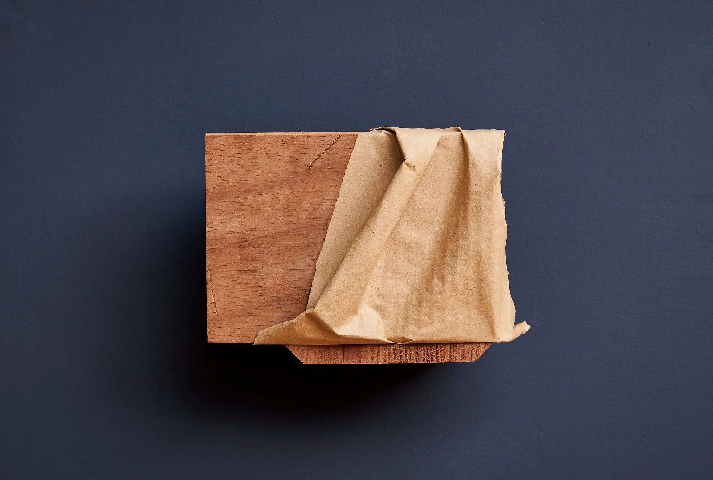 A wooden block with a brown paper bag draped over one side, set against a dark blue wall.