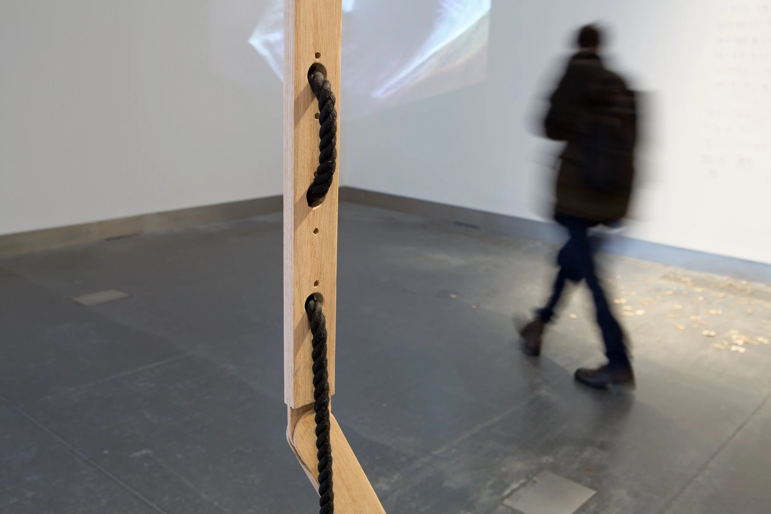 Close-up of a wooden structure with a black rope, in an indoor space with a grey floor and a blurry person walking in the background.