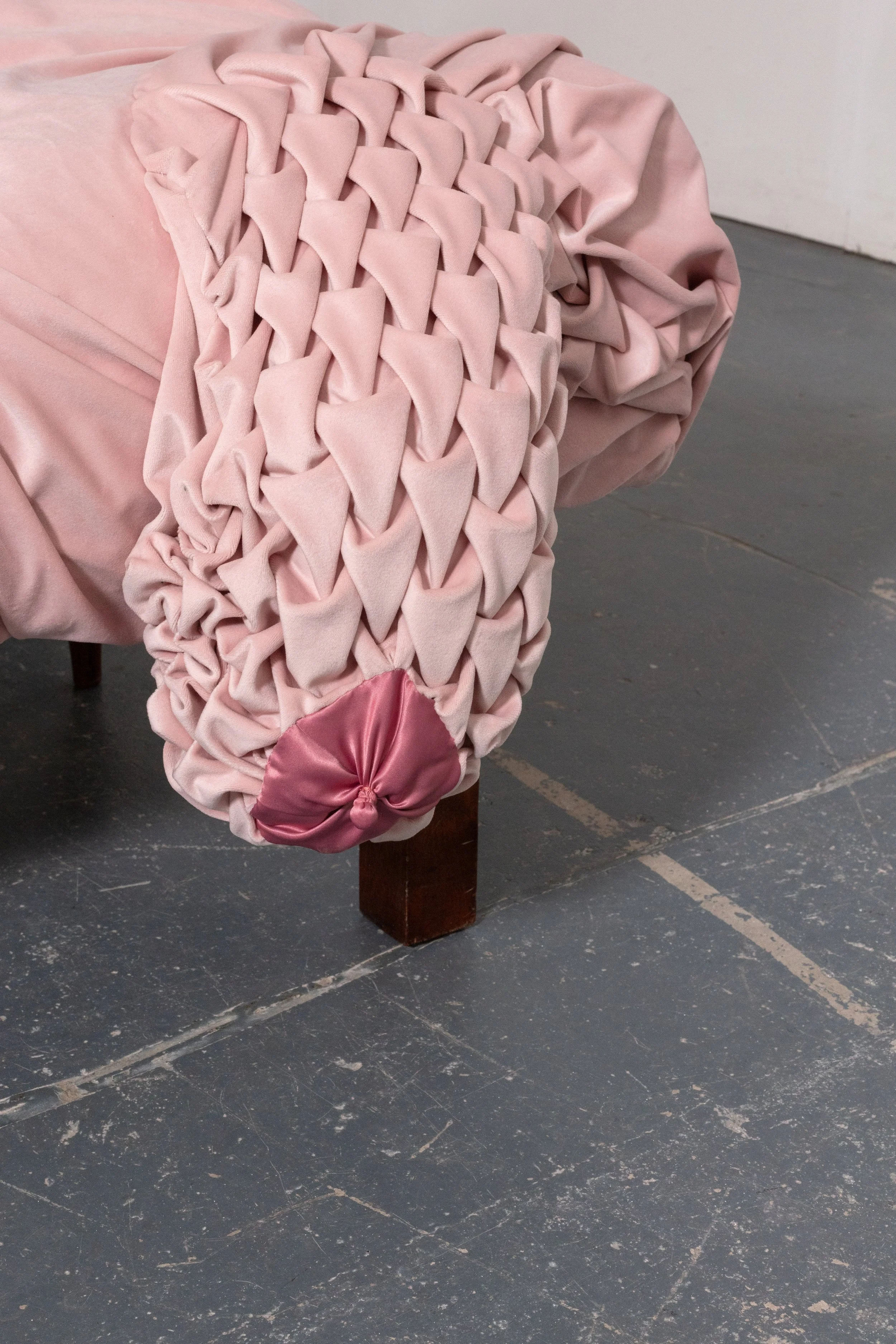 A pink upholstered chair with a decorative woven backrest and a satin pink bow at the bottom, placed on a dark gray floor.