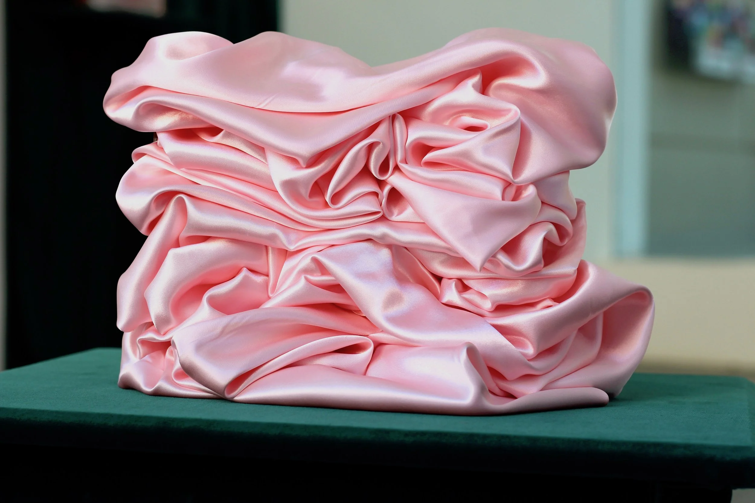 Pink satin fabric arranged in ruffles or folds on a green surface with a blurred background.