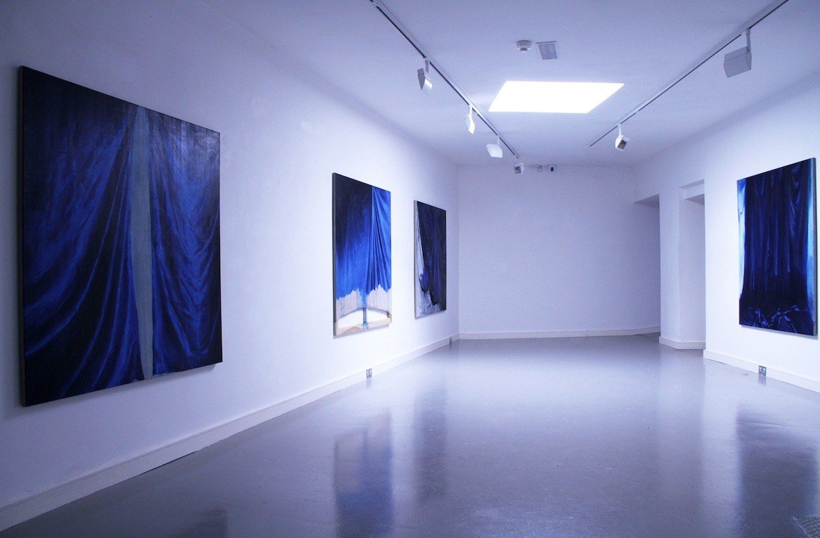 An art gallery with five abstract paintings featuring shades of blue and black, displayed on white walls under track lighting.